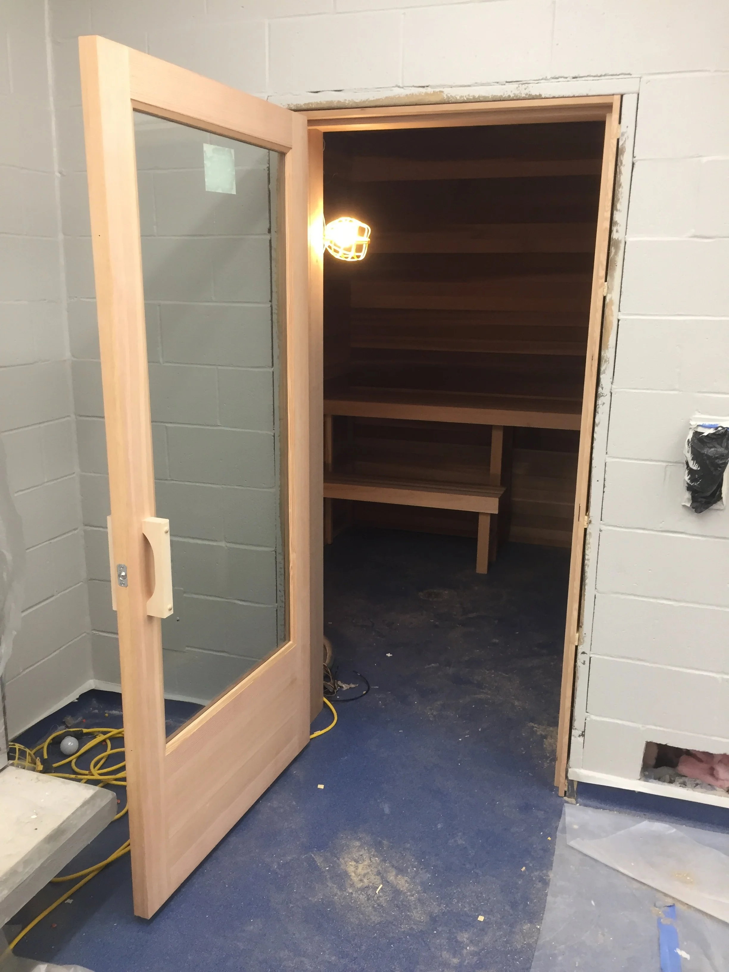 Construction site showing a partially installed wooden door with a glass window, leading into a room with dark wooden shelves and a black floor. Construction tools and electrical cords are on the floor.