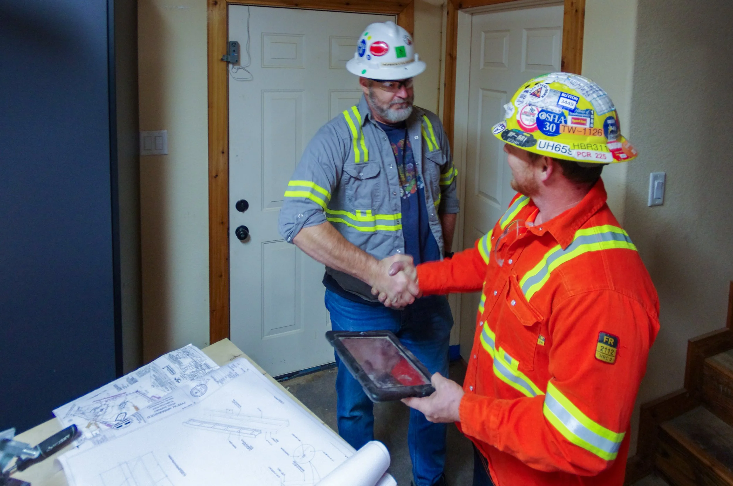Two construction workers shaking hands inside a room, wearing safety helmets and reflective clothing. One worker is holding a tablet. There are construction blueprints and tools on the table in front of them.