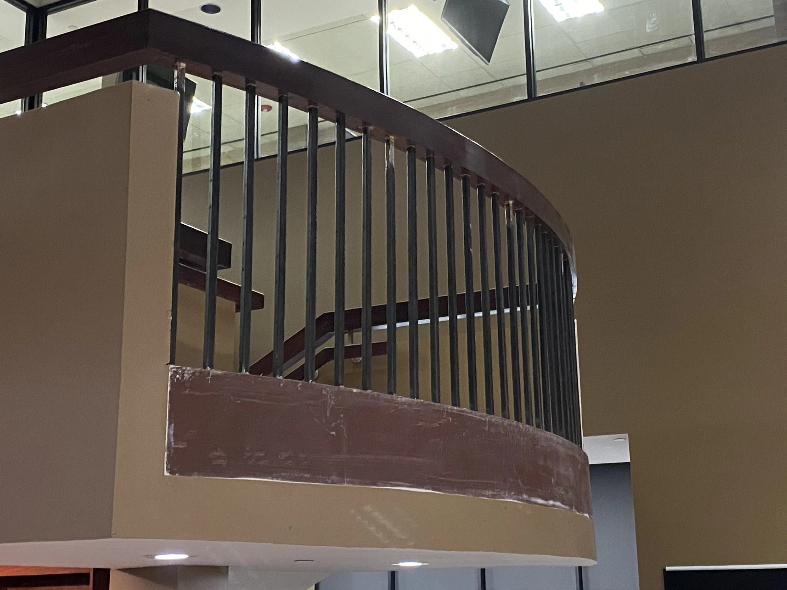 A curved staircase with wooden handrail and black metal balusters, seen from below, with a glass-walled upper level and ceiling lights.