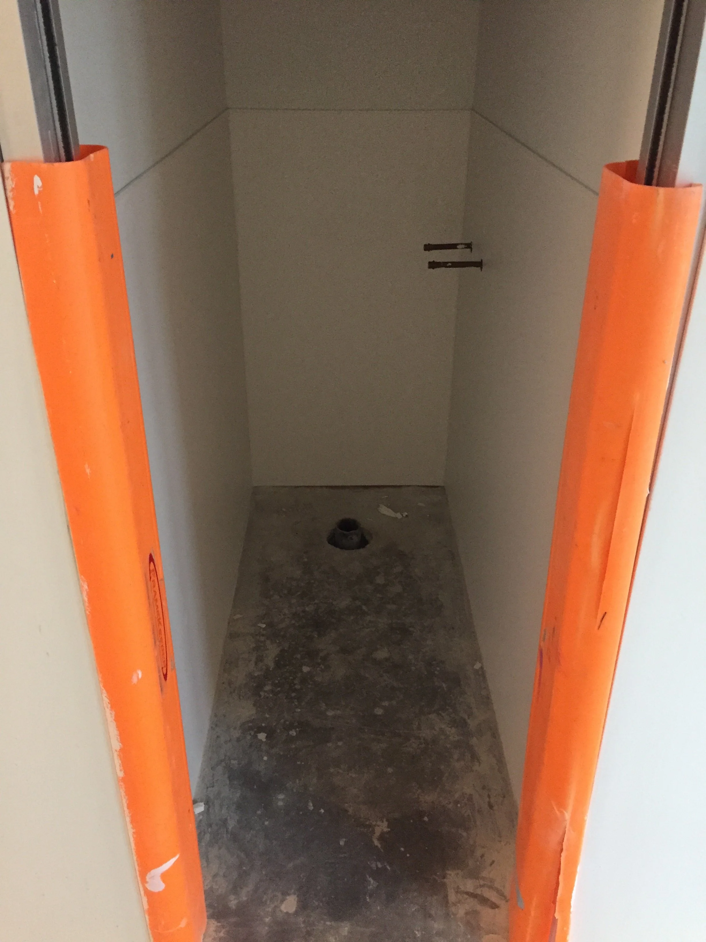 Inside a construction elevator shaft with orange safety padding on the sides, showing a concrete floor with debris and a pipe opening at the back.
