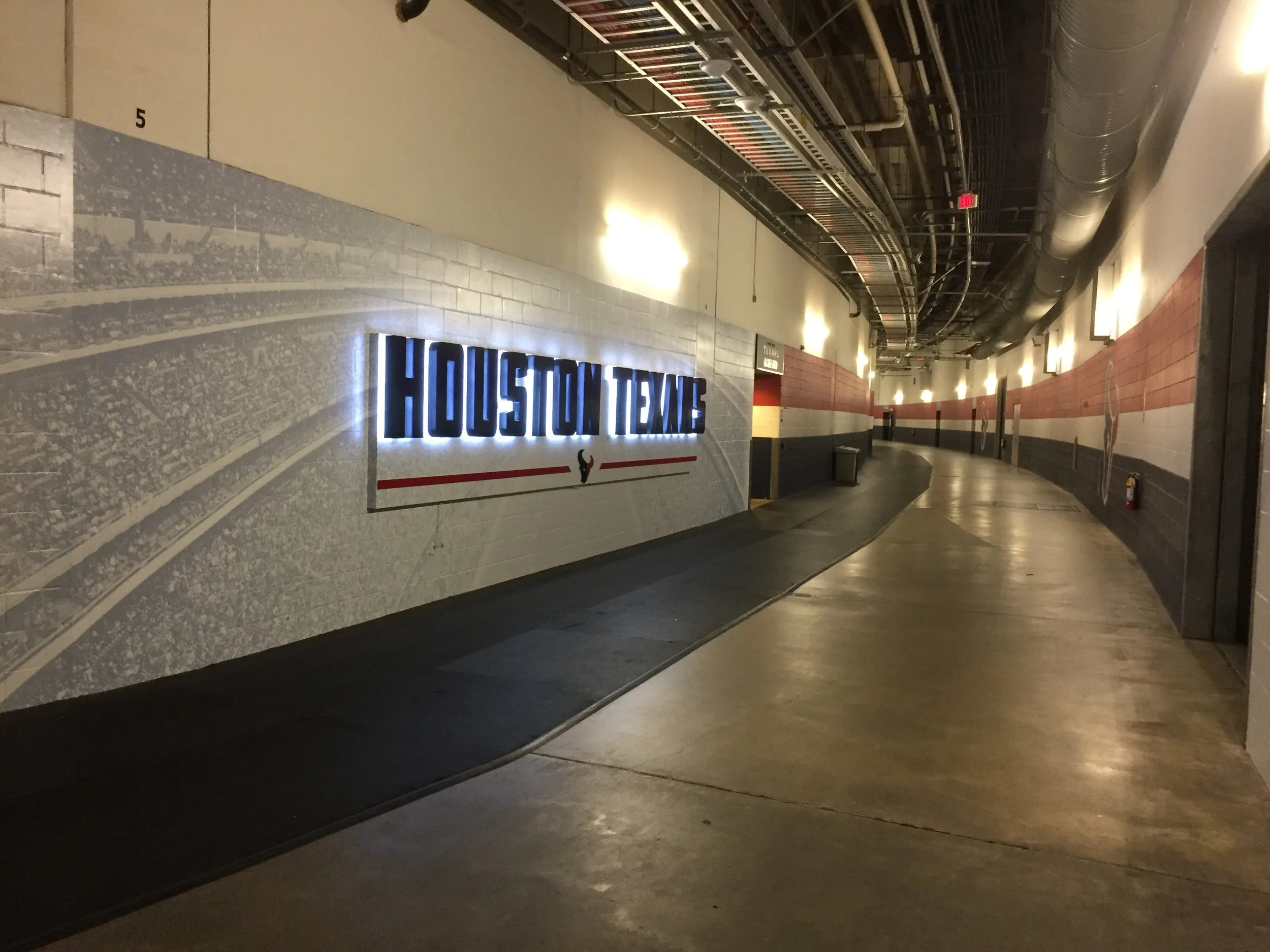 An indoor hallway with a wall displaying the Houston Texans logo, dim lighting, and a ceiling with exposed HVAC ducts and pipes.