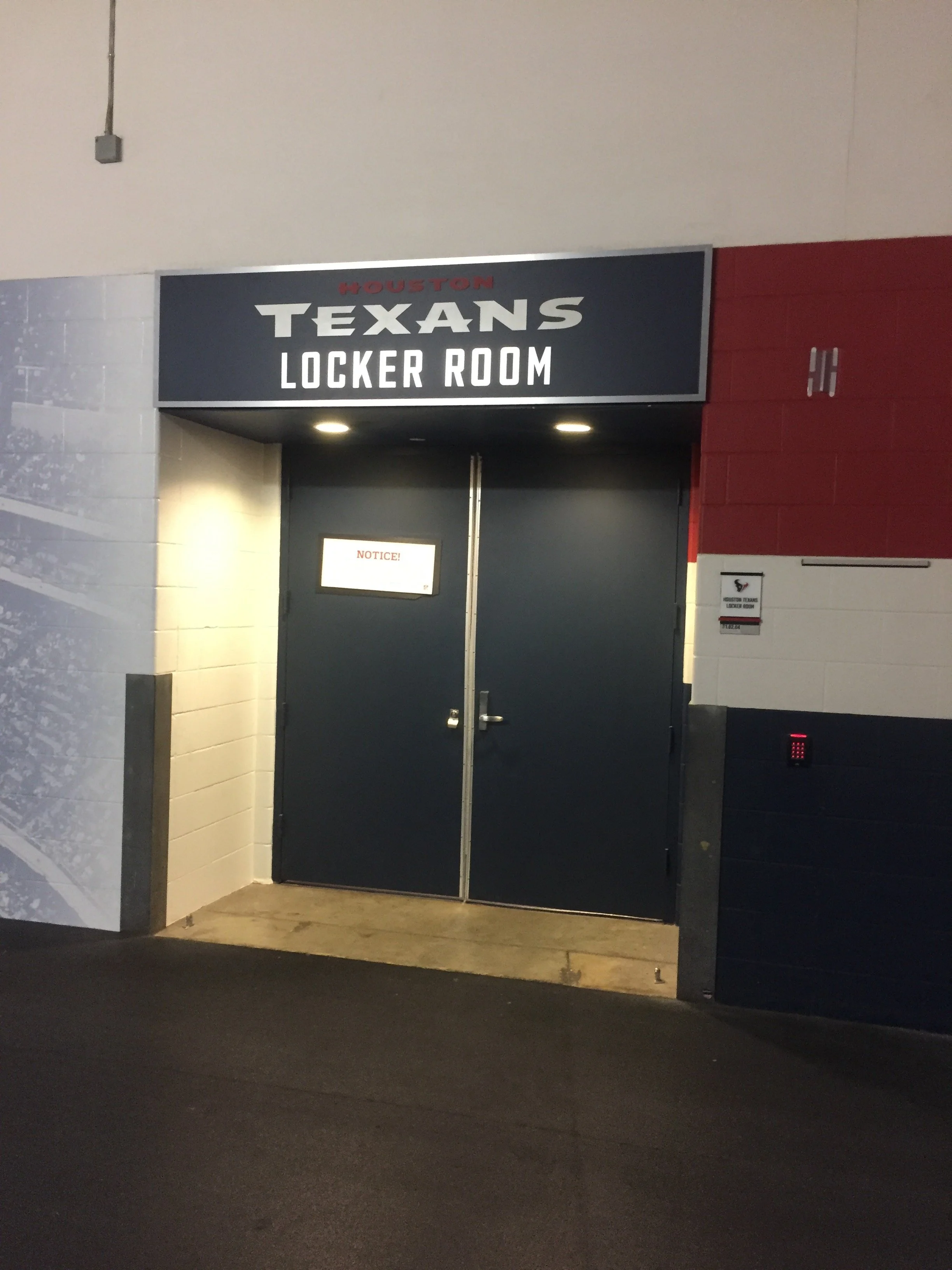 Entrance door to the Houston Texans locker room with a sign above it and a notice on the door. The wall to the right is red and white tiles, and the wall to the left is white brick.