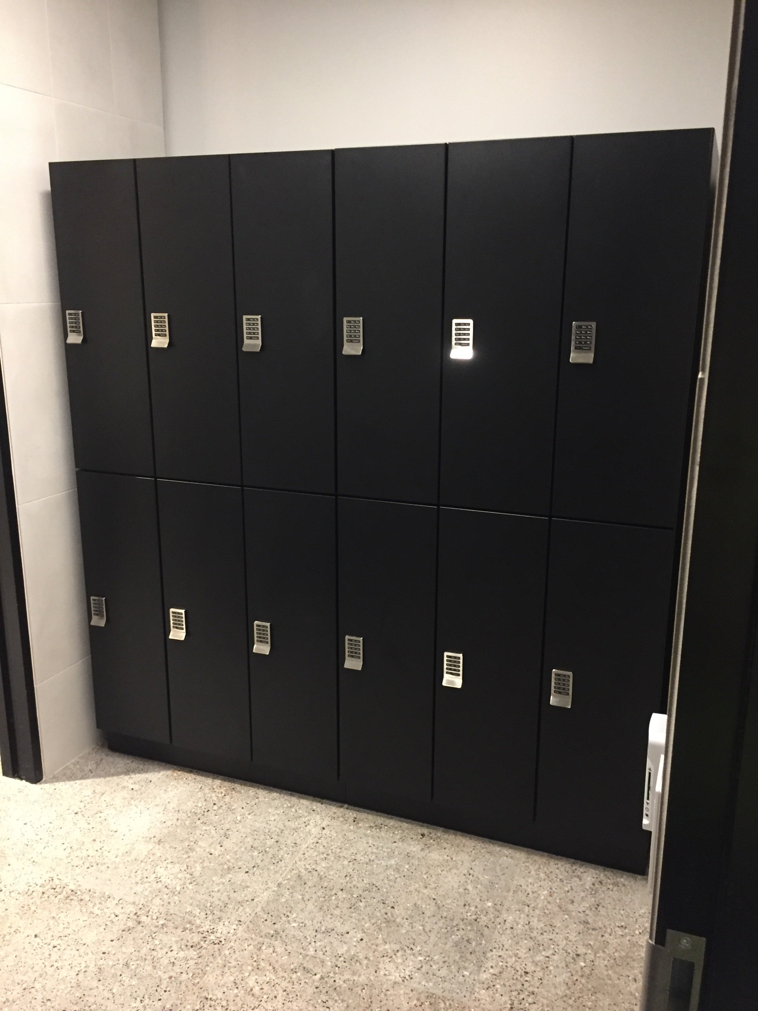 A row of black lockers with silver combination locks in a hallway with beige tiled walls and neutral gray terrazzo flooring.