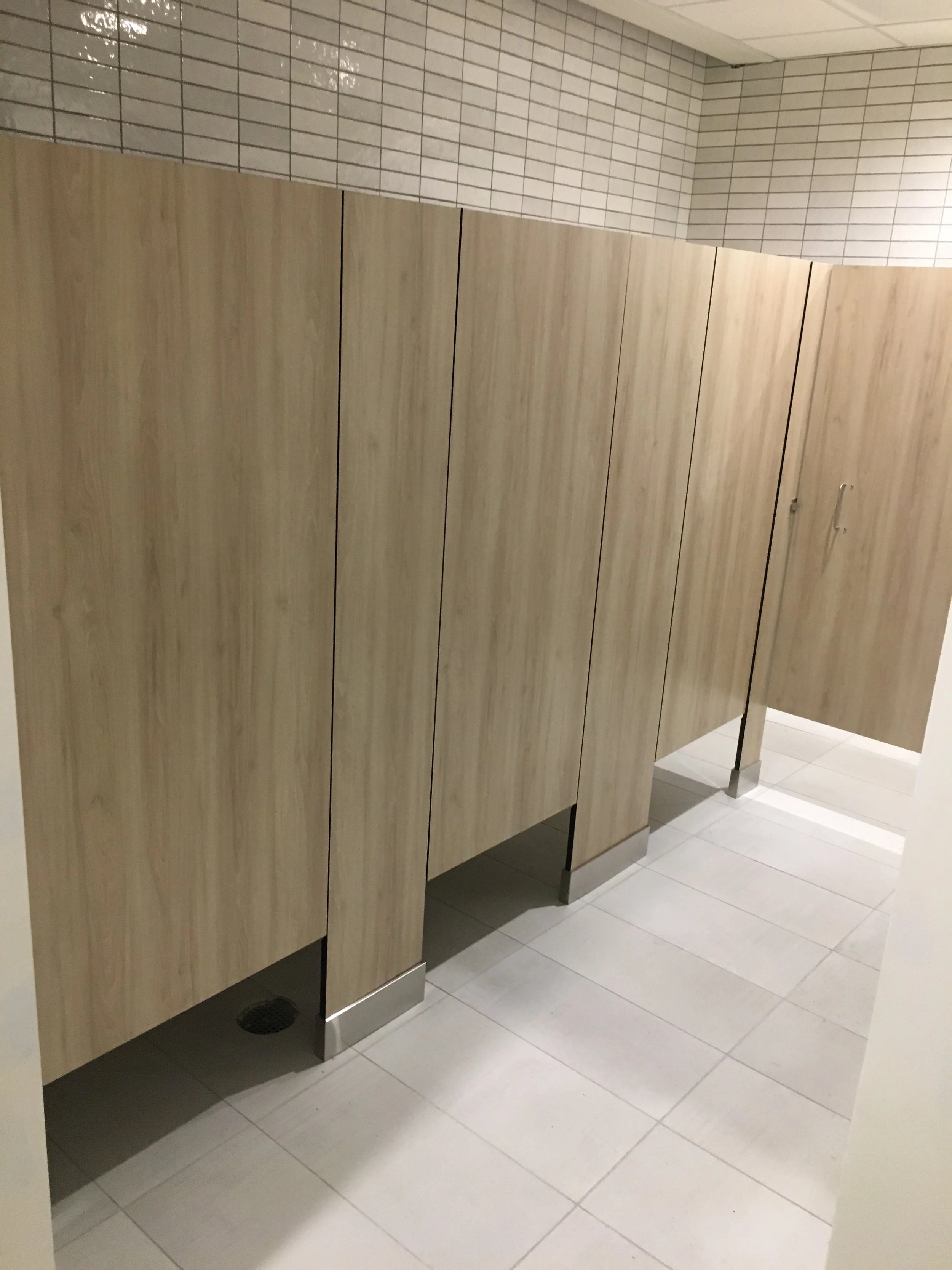Public restroom with wooden stall doors, white tiled walls, and light-colored tiled floor.