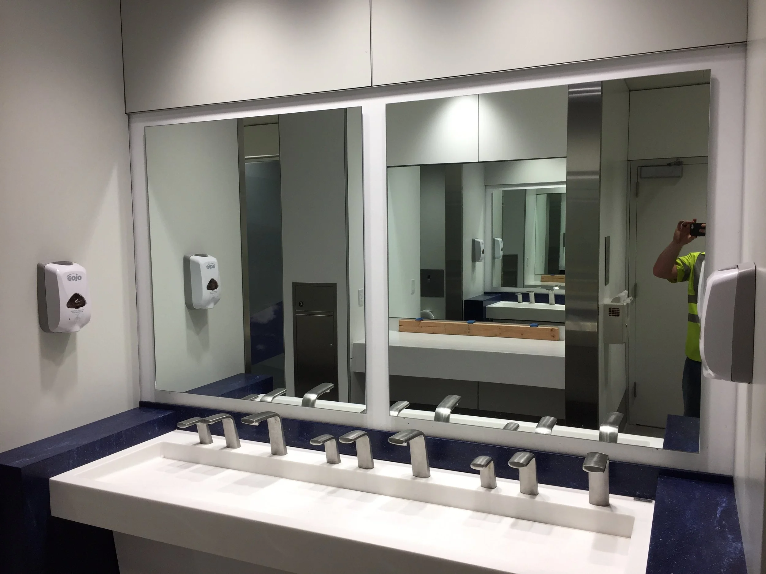 Public restroom sink area with a large mirror, soap dispensers, and sinks with soap nozzles, with a person in a high-visibility shirt taking the photo