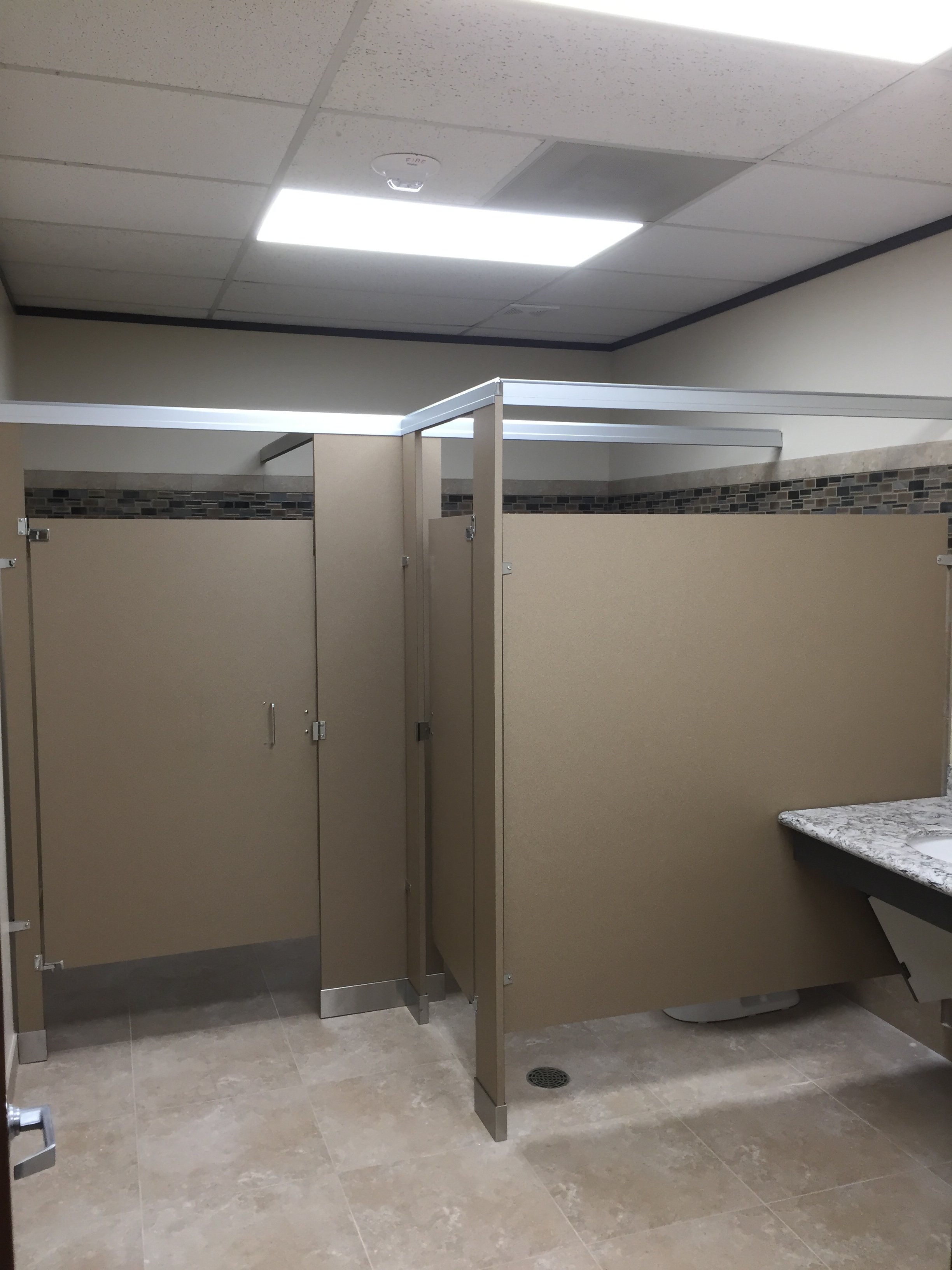 Public restroom stall with beige partition walls, a tiled backsplash with a mosaic border, a marble countertop, and tiled floor.