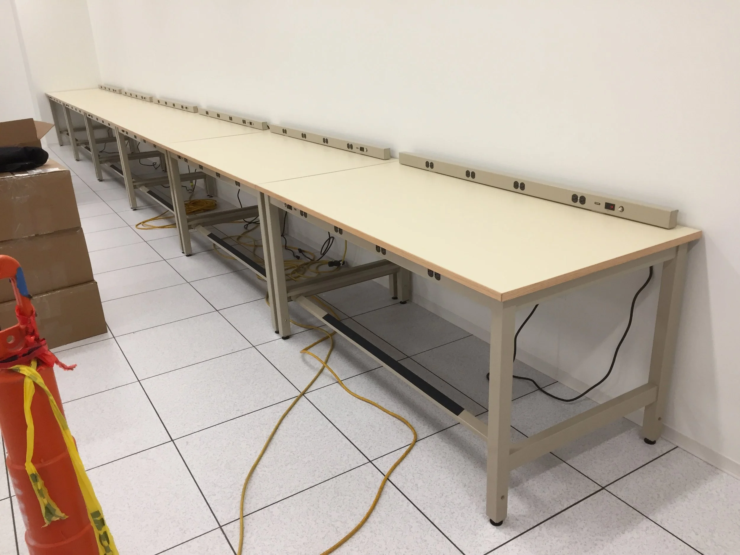Empty workstations with power outlets and cords on a long white table against a white wall in a room with tiled floor.