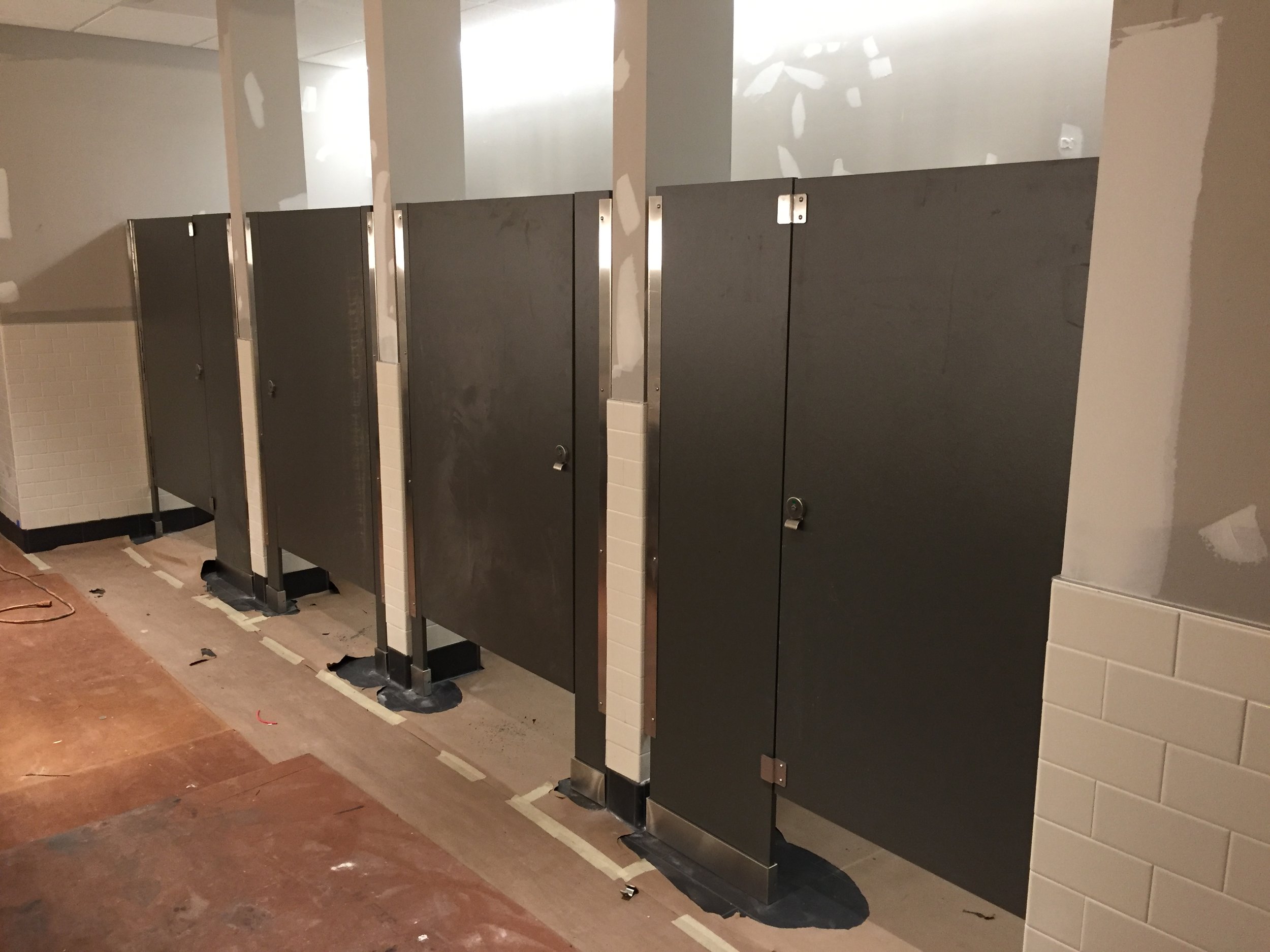 Public bathroom with a row of black toilet stall doors, some doors are missing or not yet installed, with visible hardware and hinges, and partial tile walls, unfinished floor, and exposed drywall.