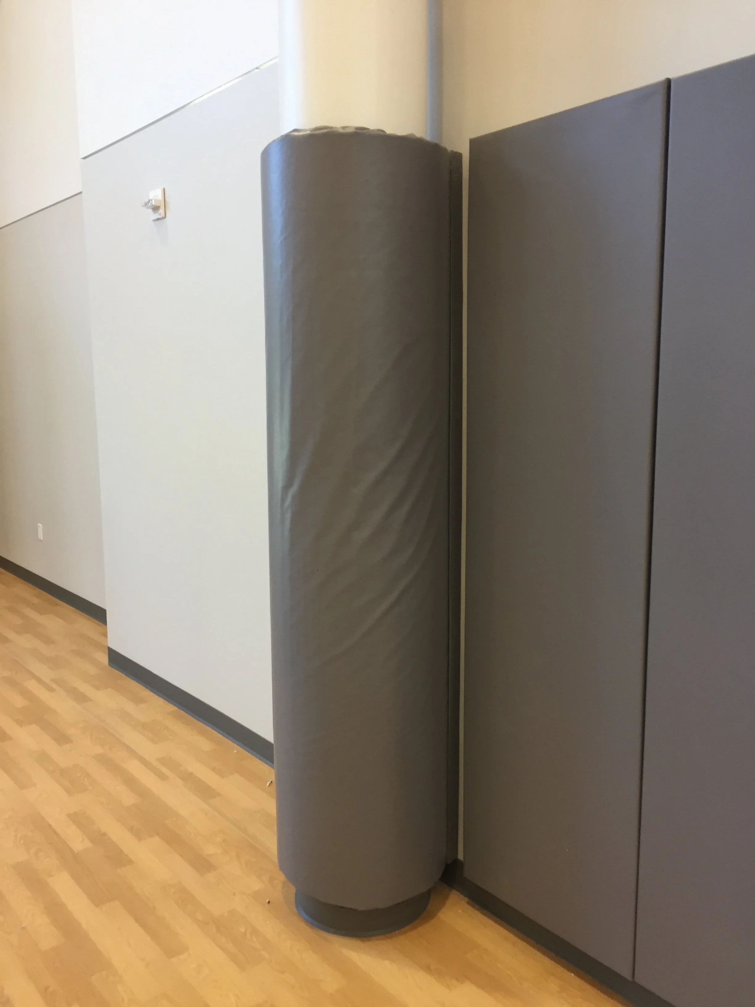 A gray, rounded object stands on a wooden floor next to a gray wall and cabinets in an indoor space.