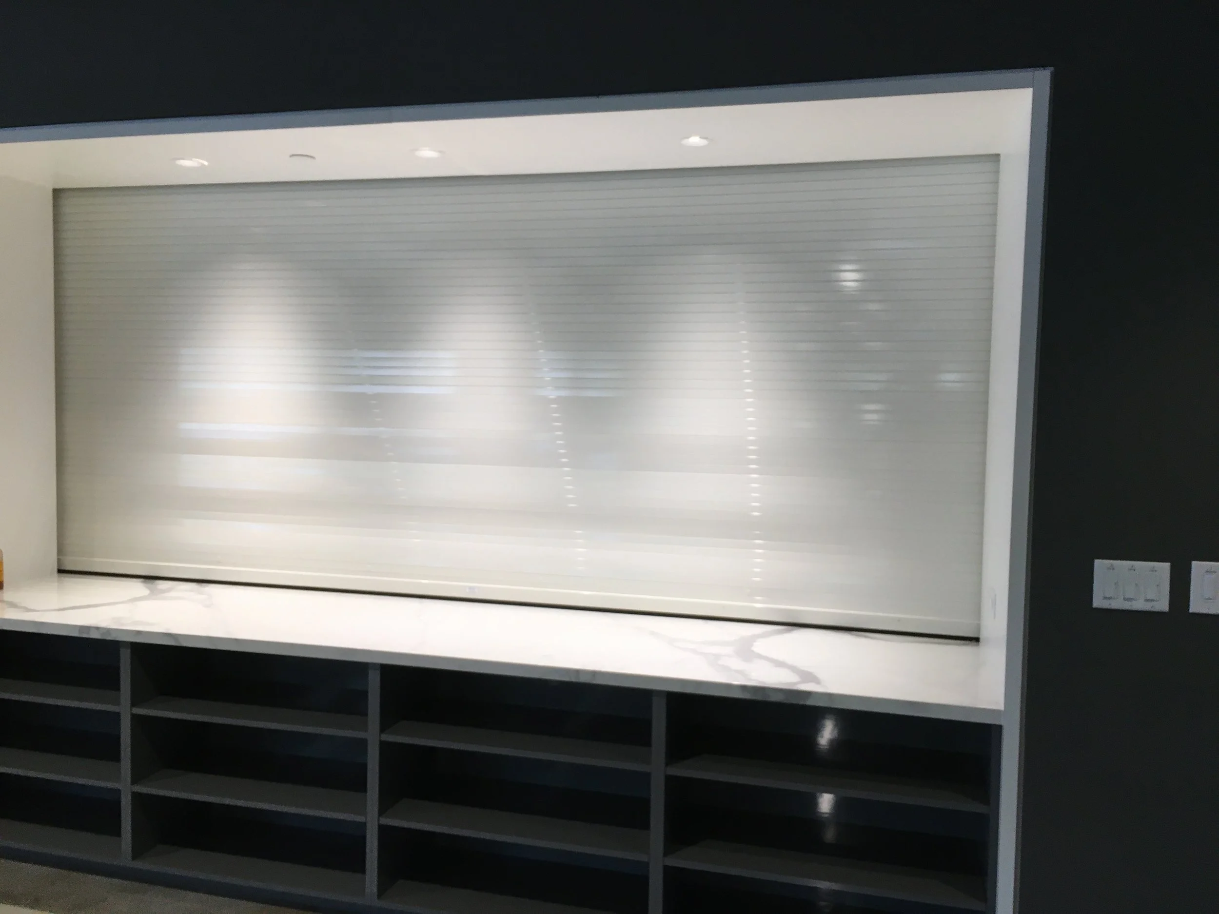 Empty retail display case with closed metal shutters, situated above open shelves, in a modern store setting.
