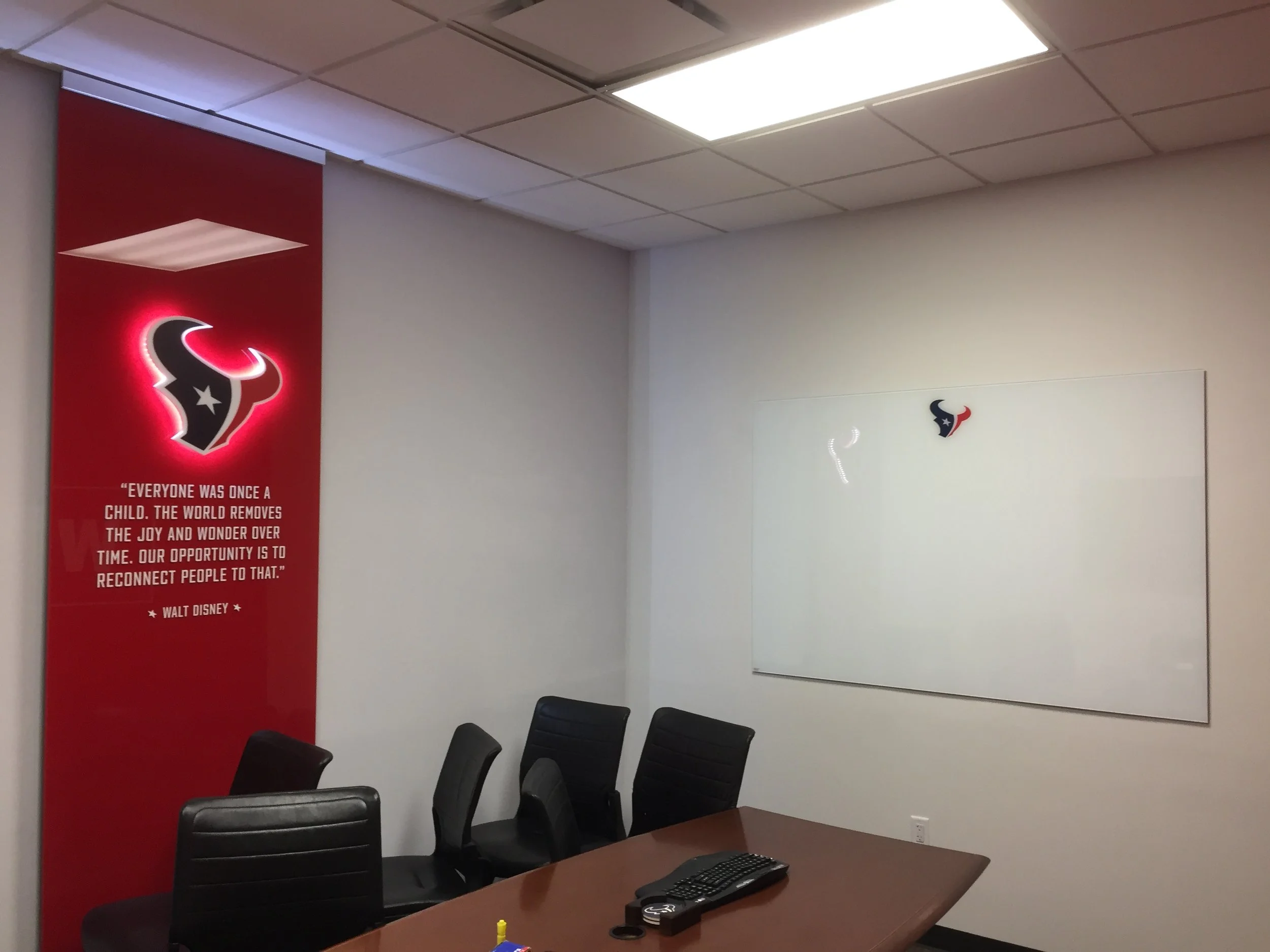 A conference room with a wooden table, black chairs, a whiteboard, a wall with a Houston Texans logo, and a red poster with a quote.