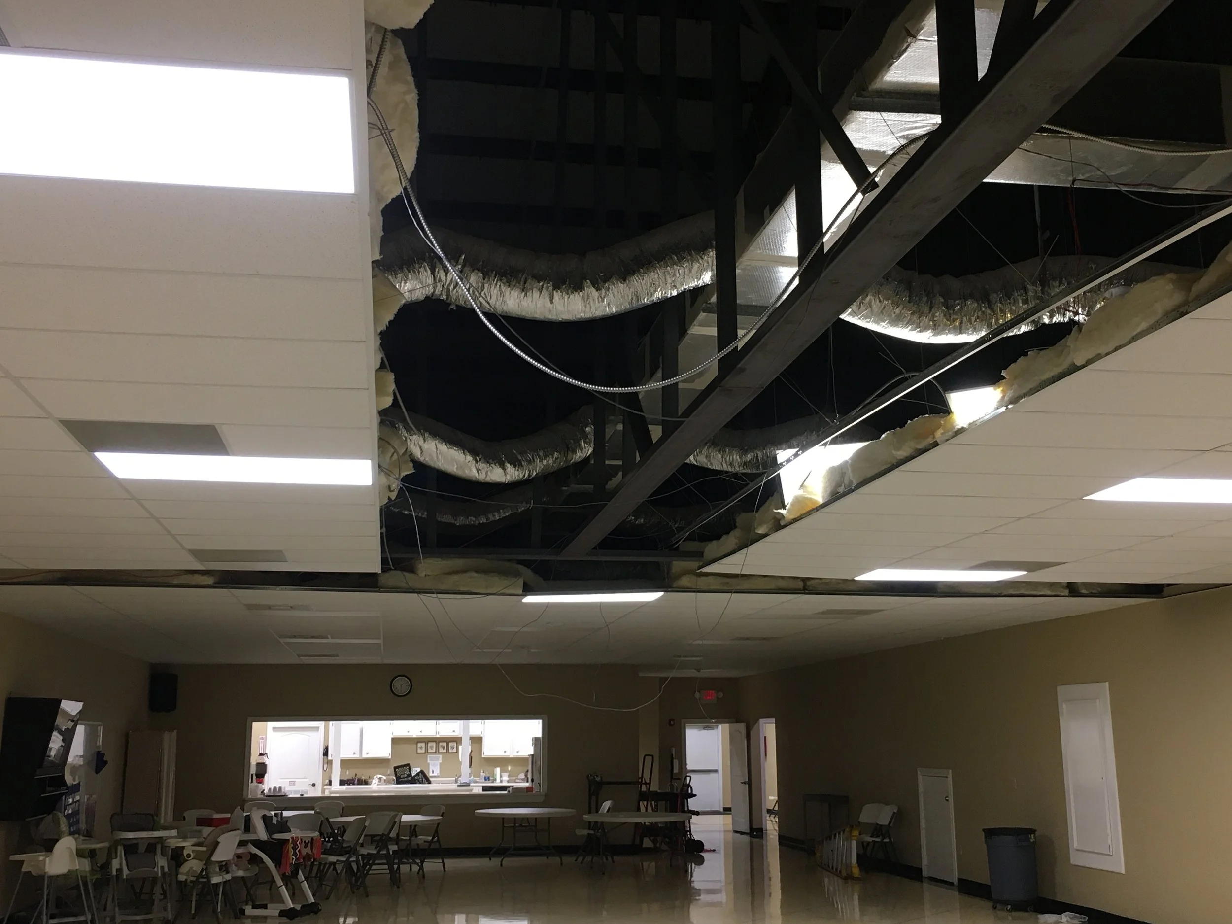 The ceiling is under construction with exposed ducts and insulation. The room below appears to be a community or meeting room with chairs, tables, and a reception area, illuminated by bright fluorescent lights.