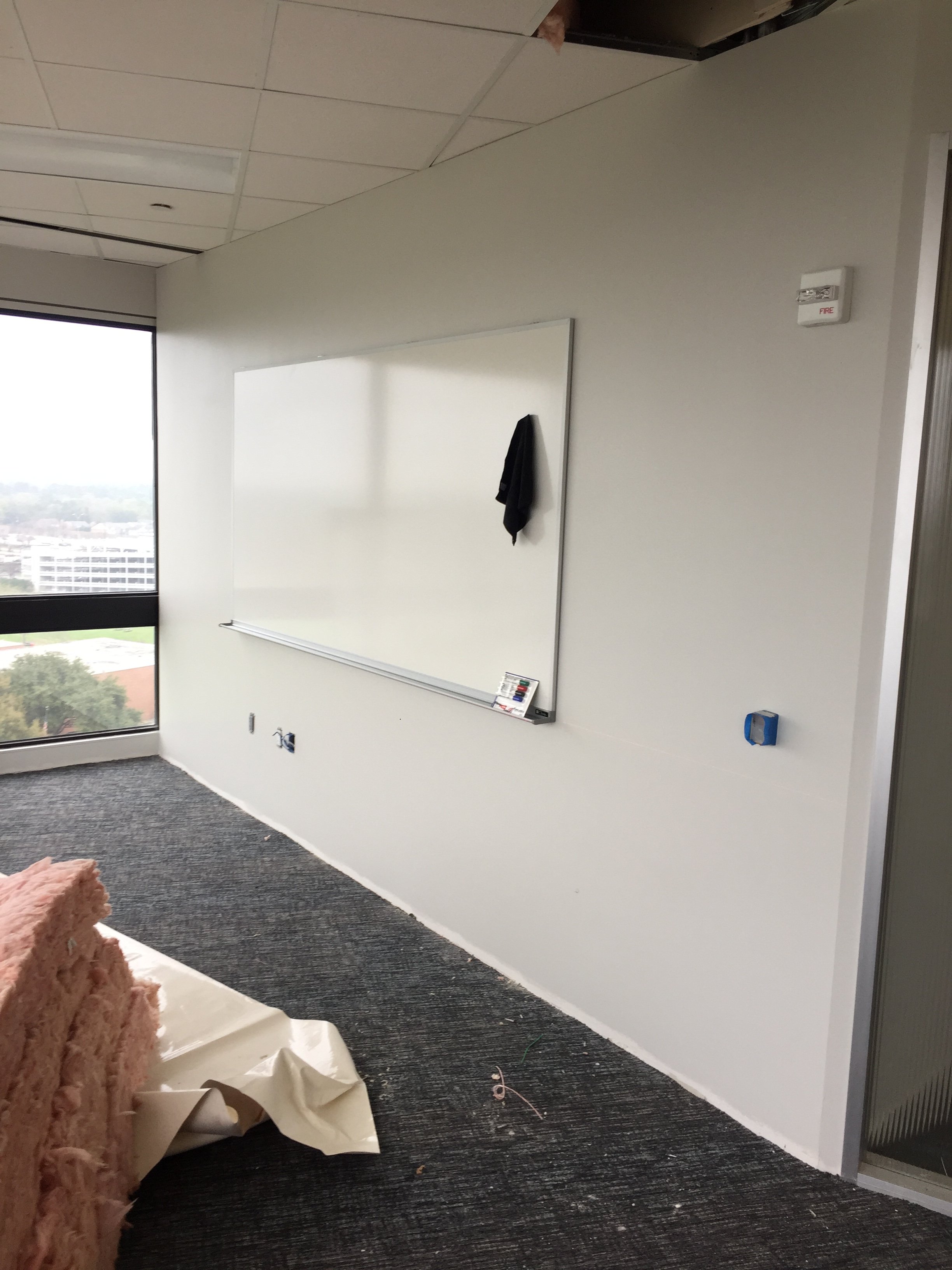 Interior of a room under construction with a white wall, a large window, a whiteboard, and construction materials on the floor.