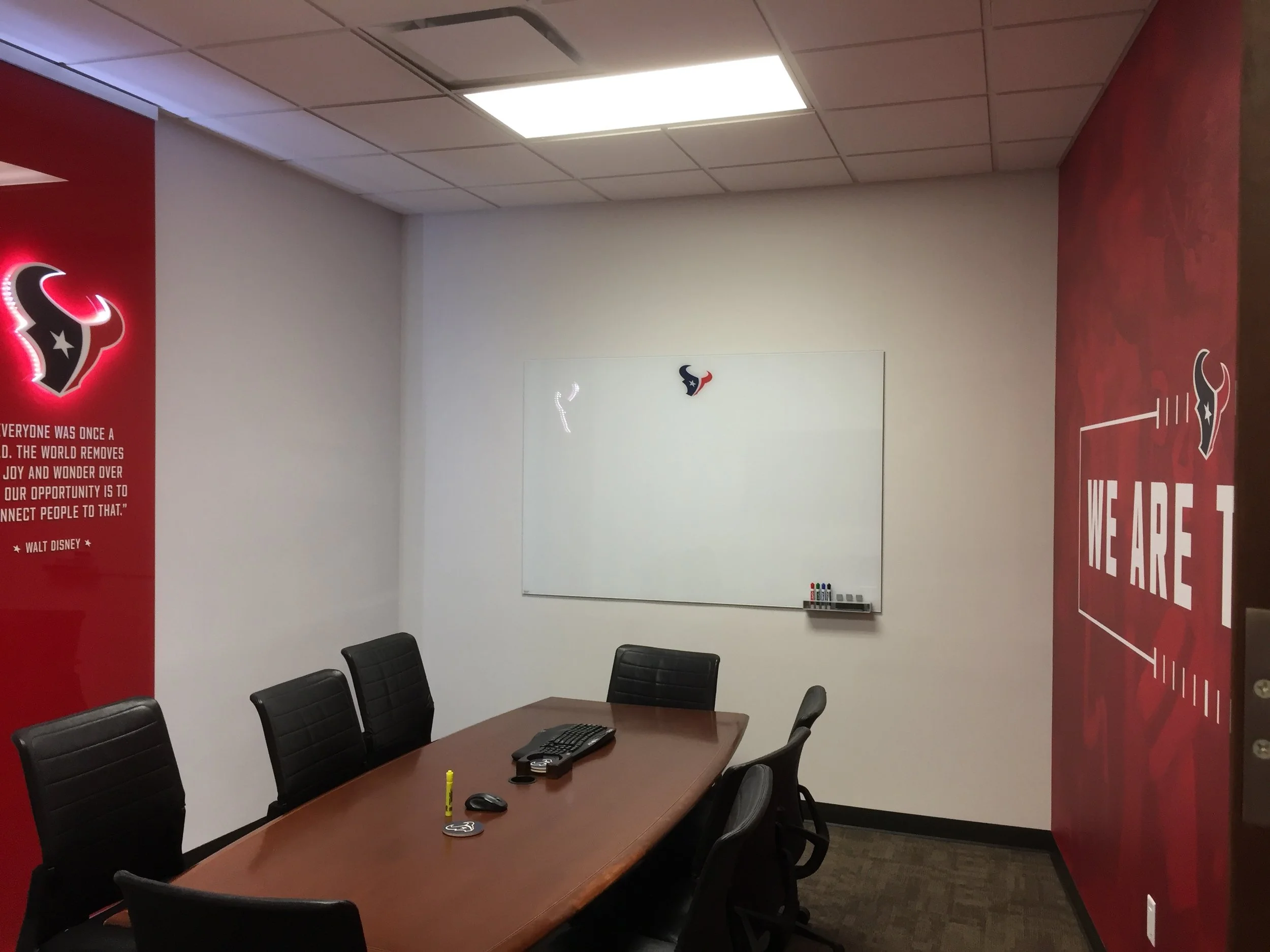 A conference room with black chairs around a wooden table. There is a whiteboard on the wall and red Houston Texans banners on the walls. The room has a ceiling with fluorescent lights.