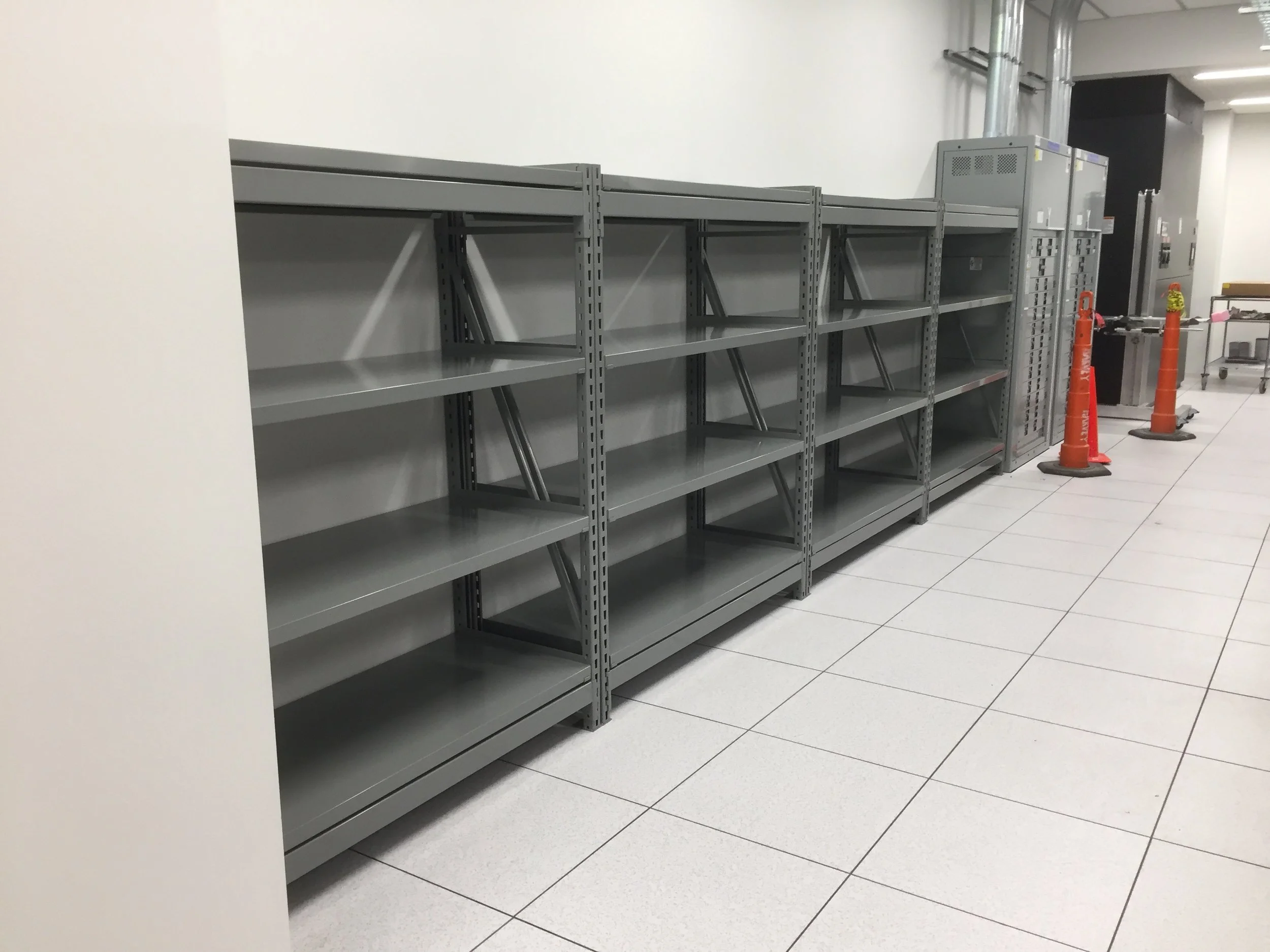 Empty metal shelving units in a white-walled room, with a black wall and some equipment in the background, and a couple of orange traffic cones on the right.