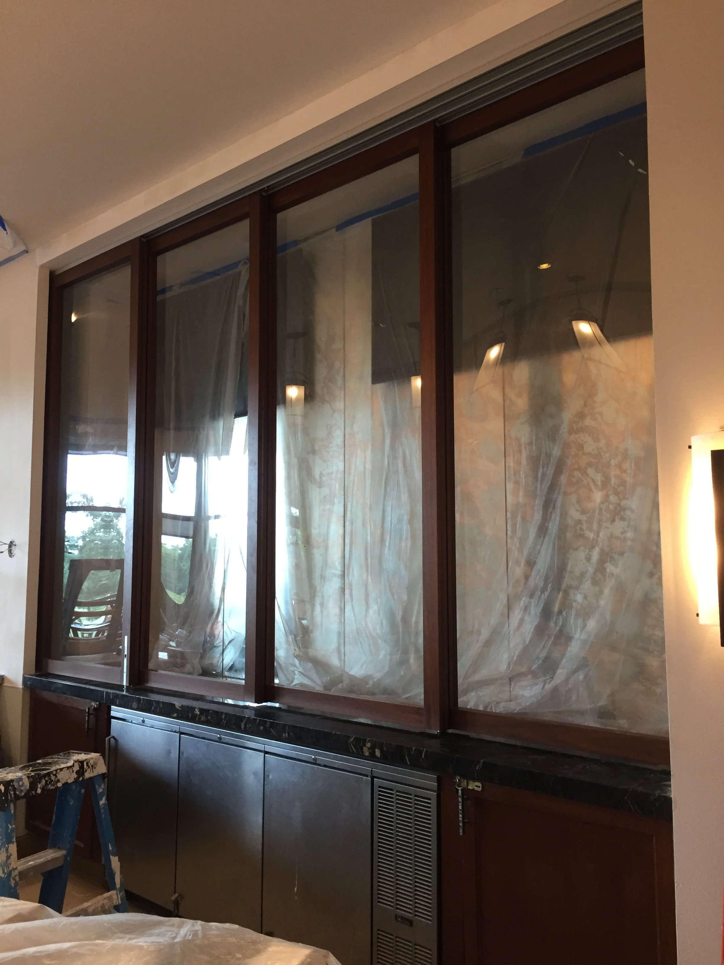 Interior of a room under renovation with a large glass window or door framed with wood, reflecting light fixtures, and covered by plastic sheeting for protection.