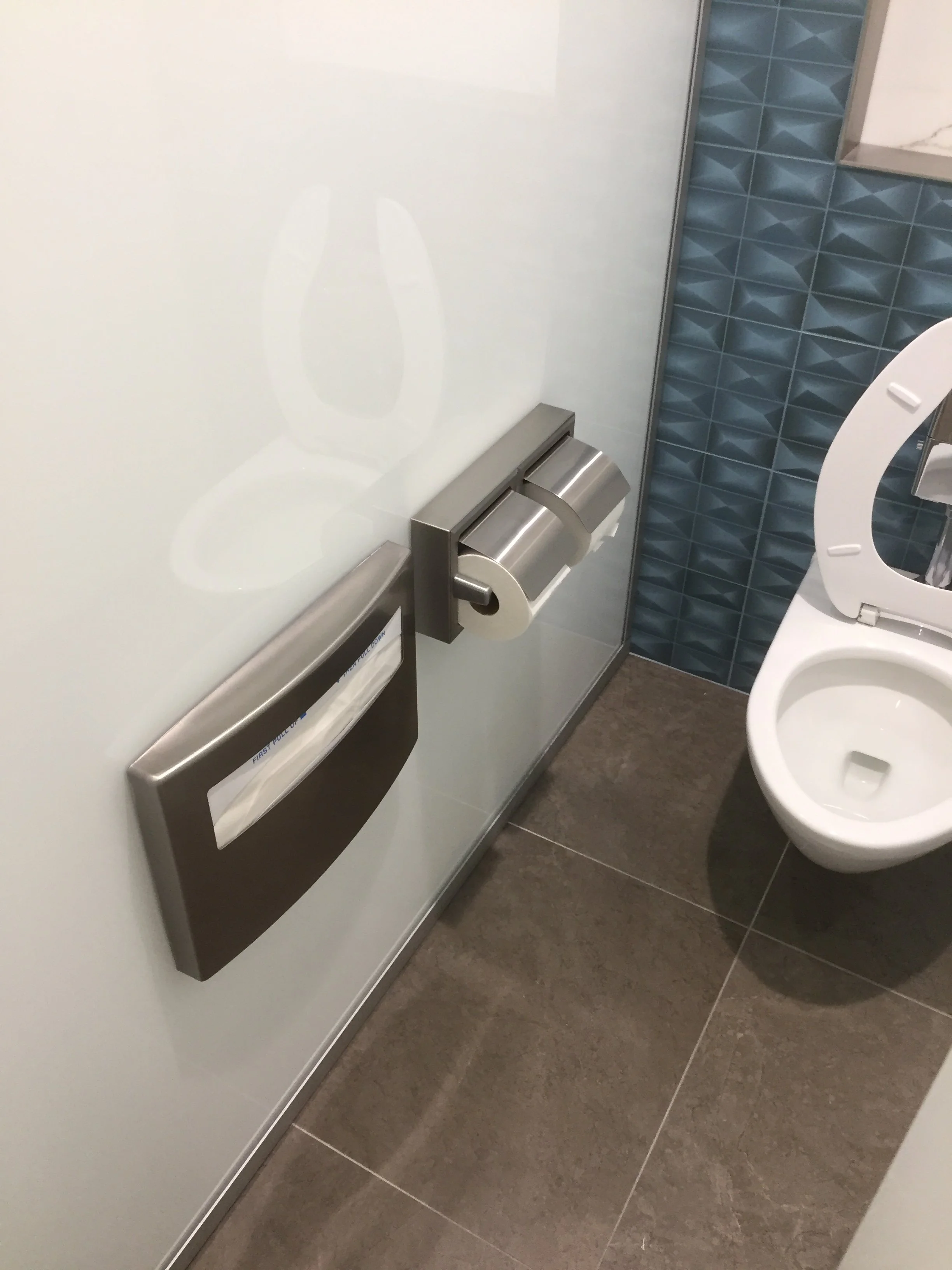 An automatic toilet with an open lid, a stainless steel toilet paper dispenser mounted on a white wall, and blue decorative tiles on the wall.