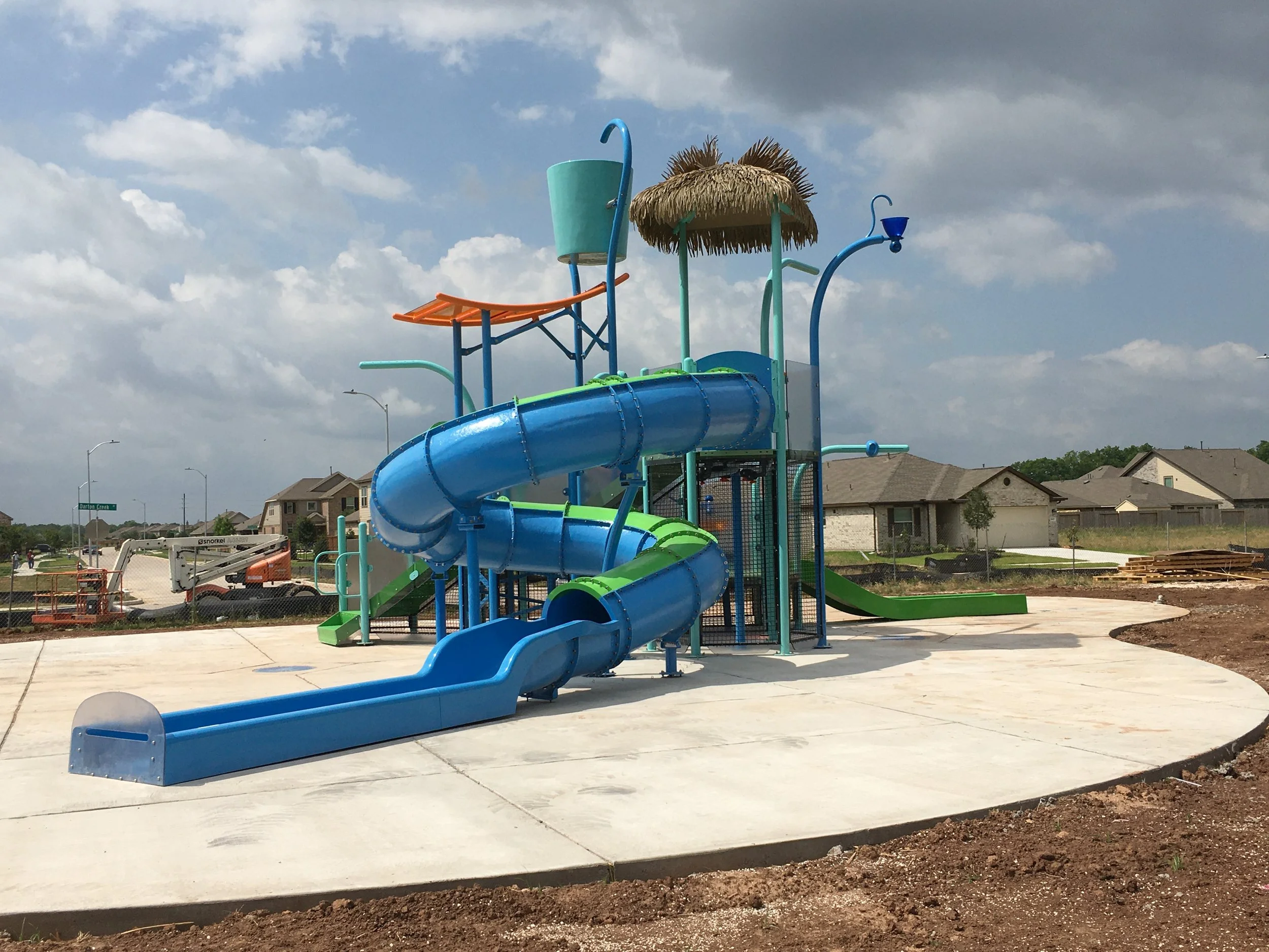 Colorful water slide at a playground under a partly cloudy sky, with residential houses in the background.