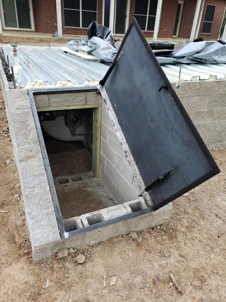 An outdoor electrical or utility access hatch with a hinged metal cover, built into a concrete base, with a plumbing or electrical pipe inside.