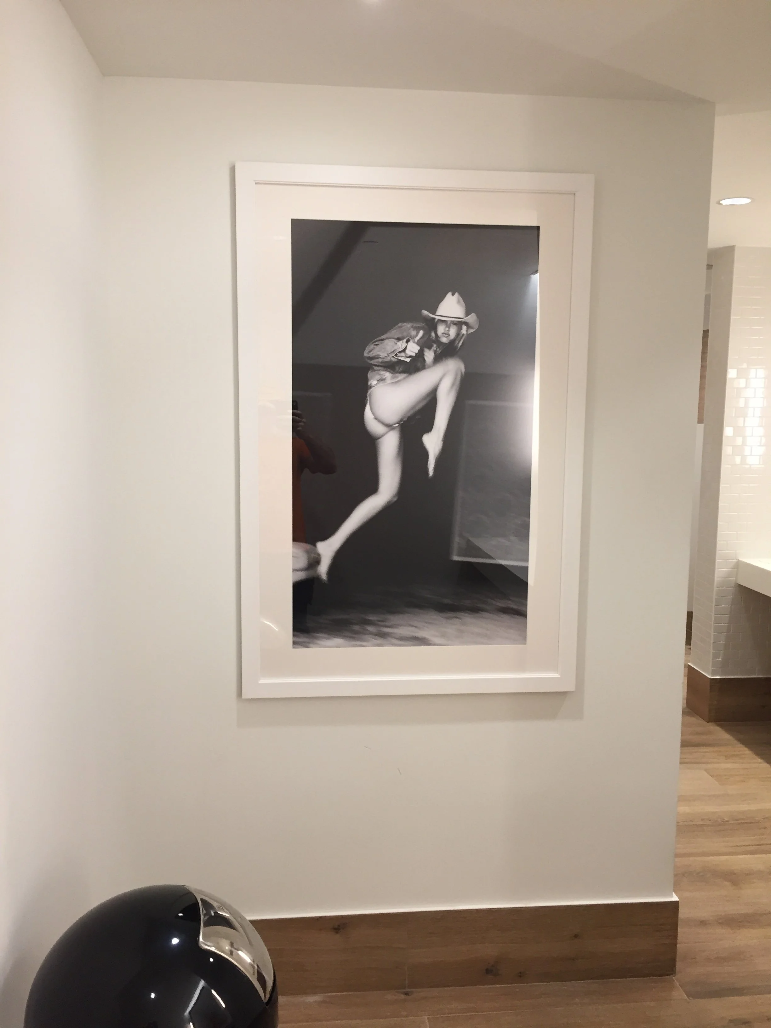 A black and white photograph of a woman wearing a cowboy hat, a jacket, and underwear, striking a pose with one leg raised and a hand on her hip, displayed on a wall in a gallery.