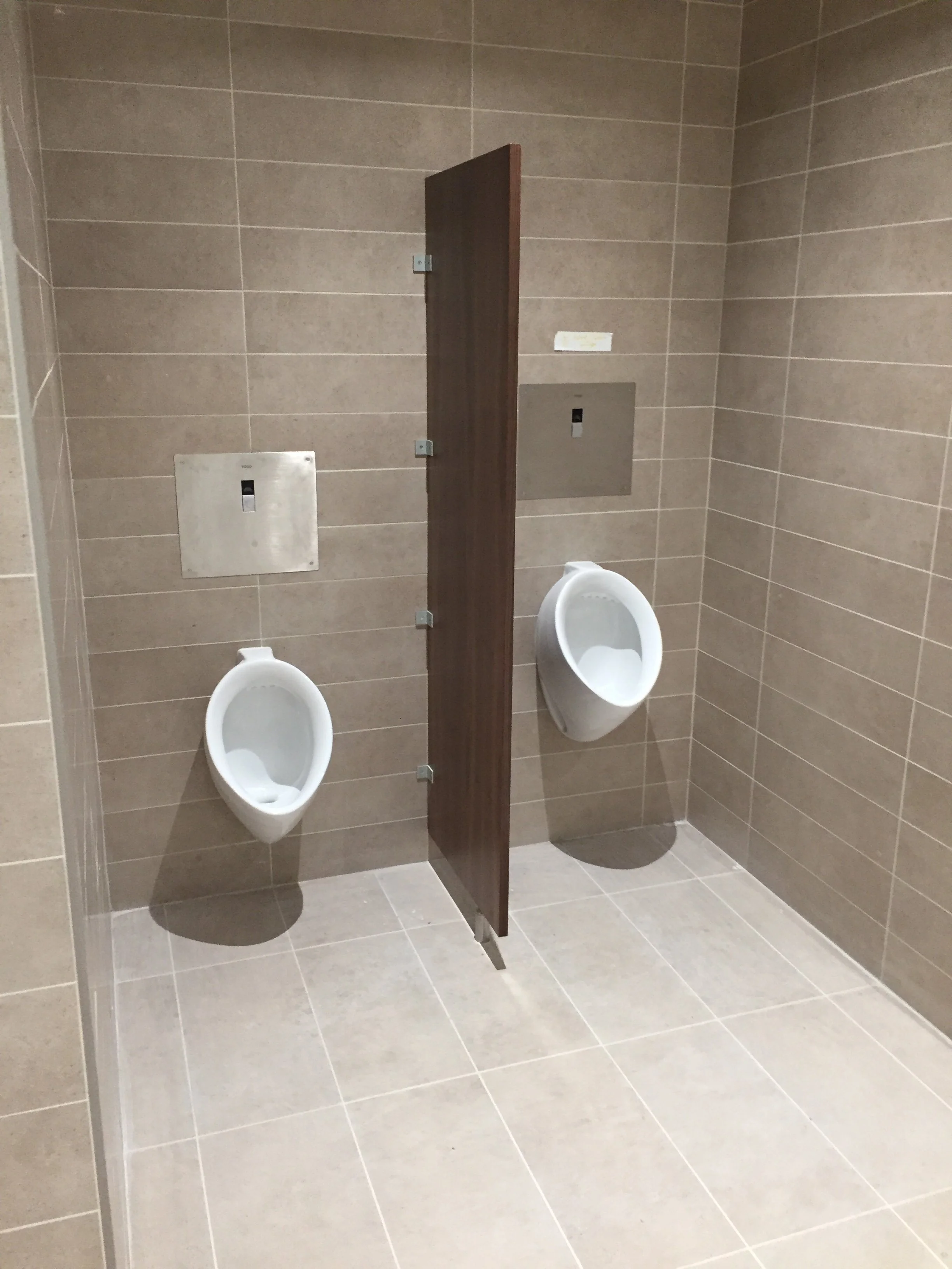 Public restroom with beige tiled walls and floor, two urinals, a wooden privacy divider, and metal access panels on the wall.