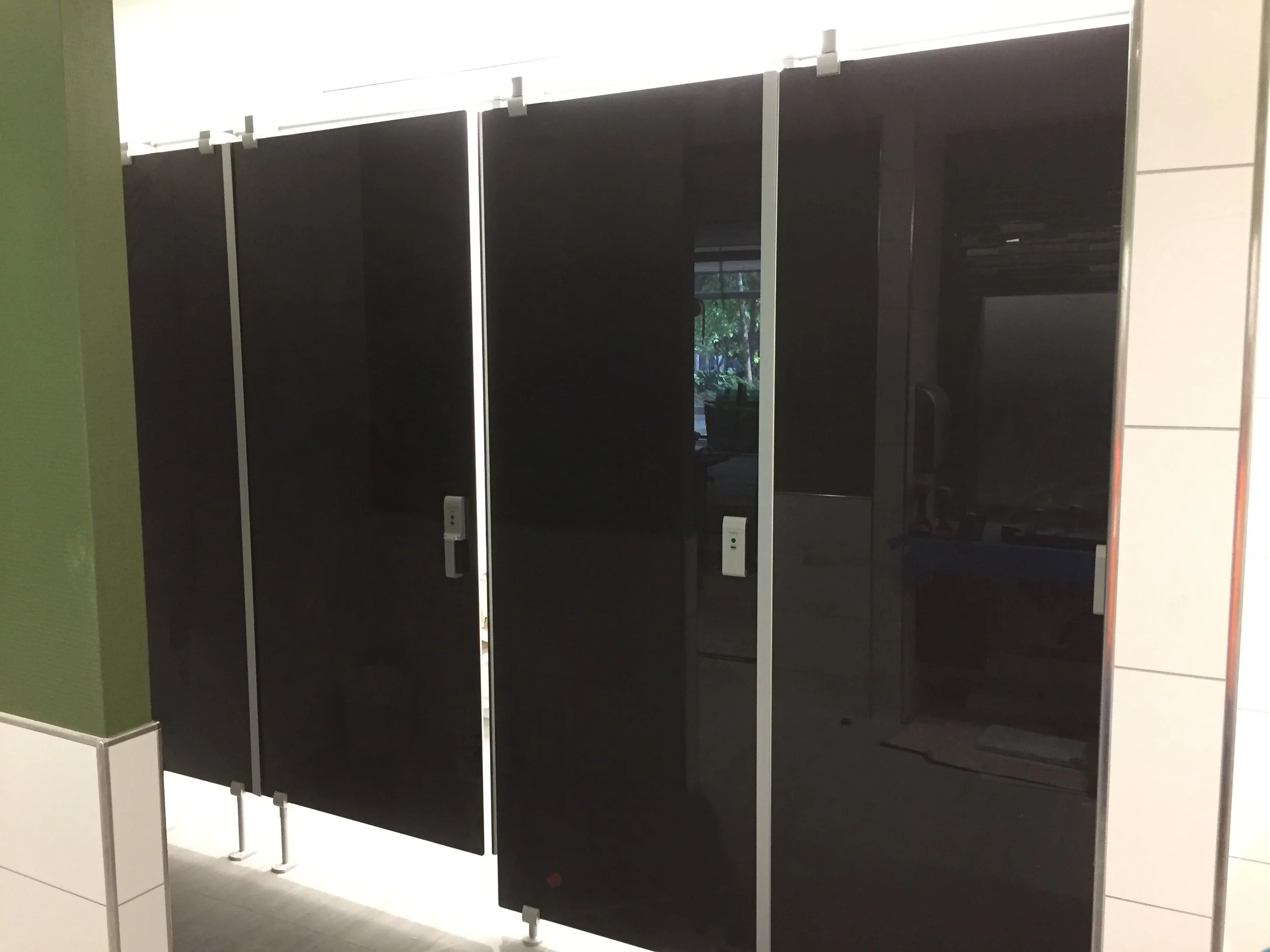 A row of black stalls in a restroom or locker room, with metal hooks at the top and electrical outlets at the bottom, against a white-tiled wall.