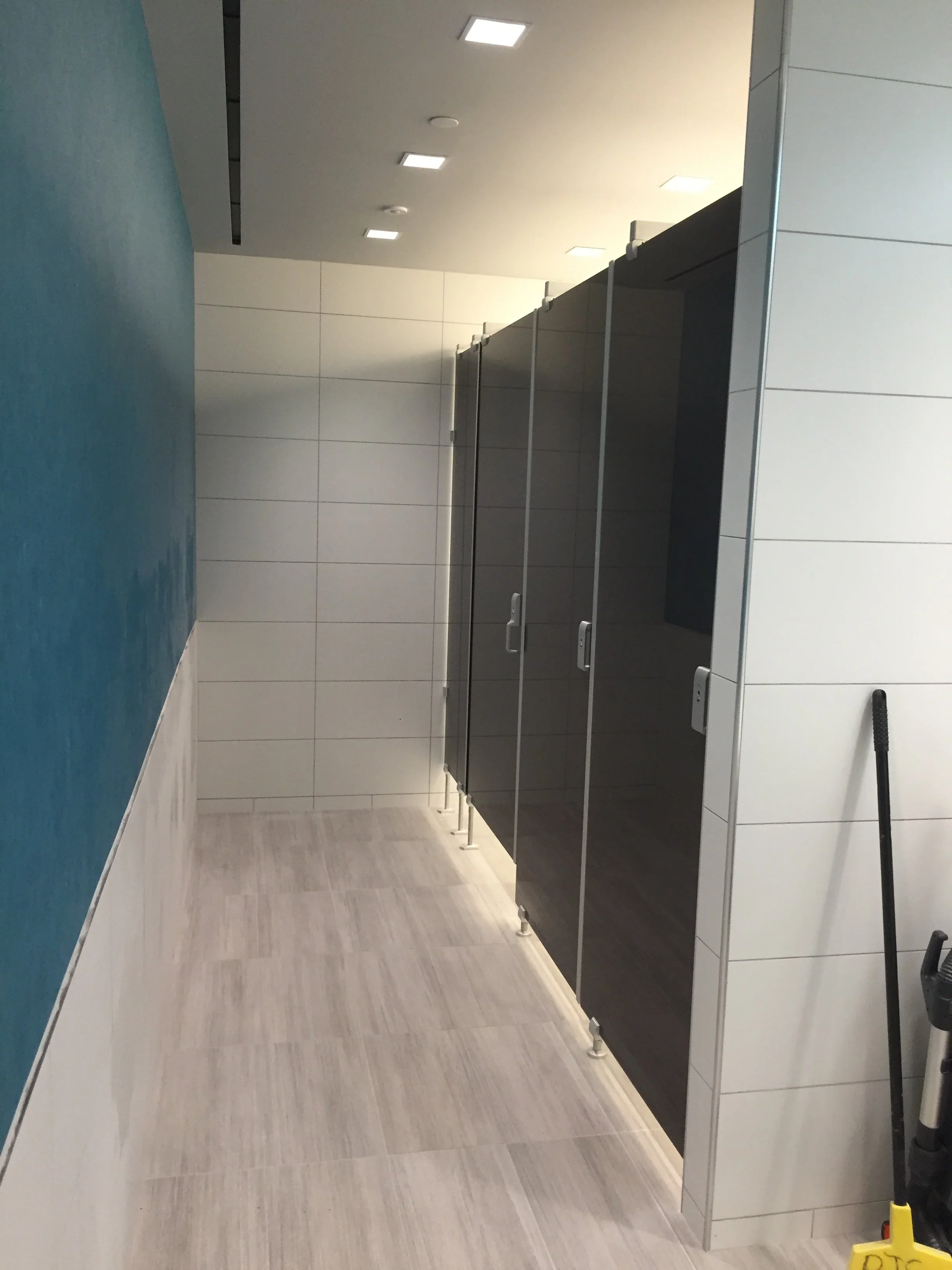 Public restroom with changing stalls, white tiled walls, wooden floor, and cleaning supplies visible on the right.