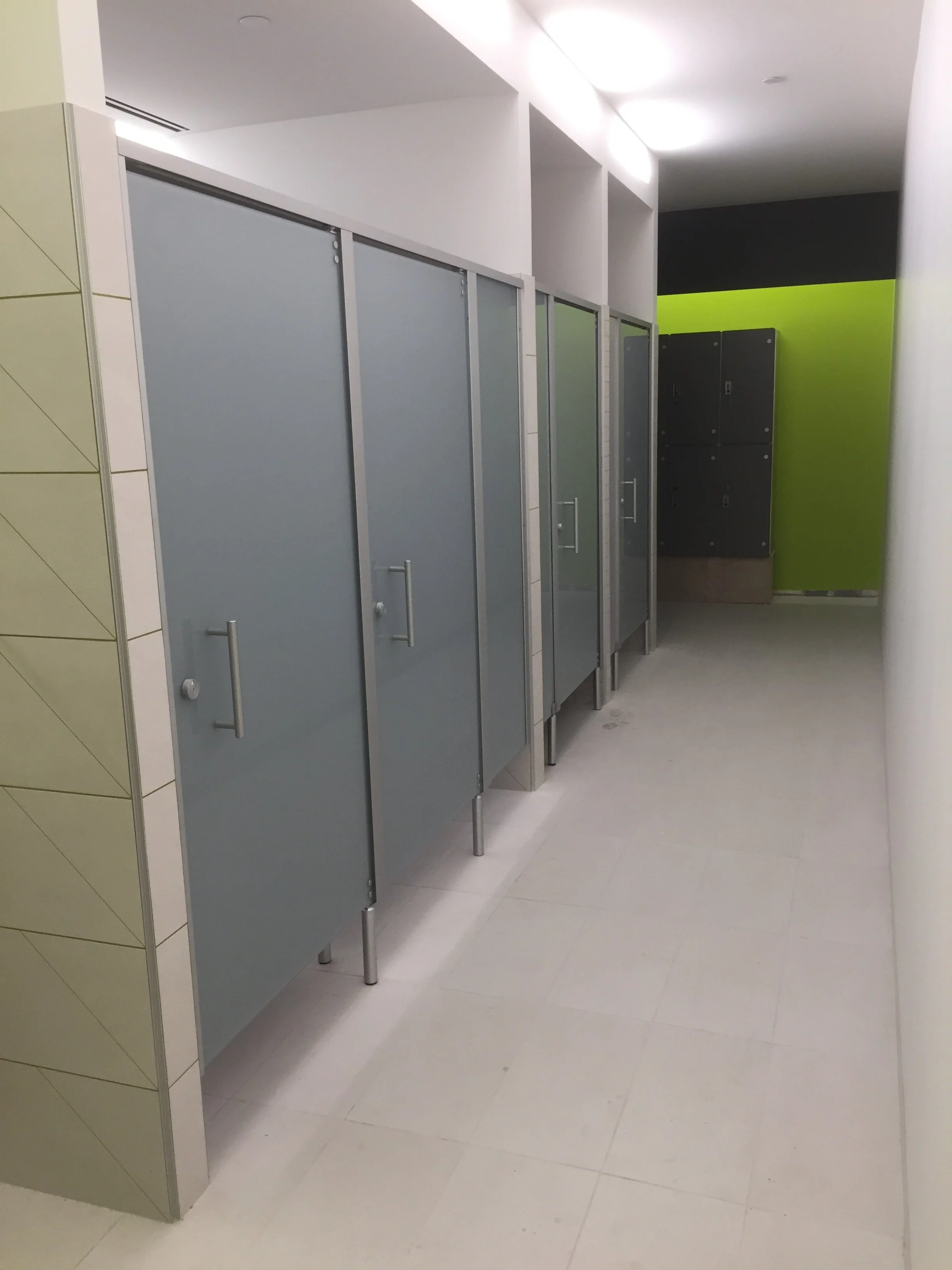 Public restroom with gray stalls, yellow tiled wall on the left, lime green wall in the back, white tiled floor, and a white ceiling with recessed lighting.
