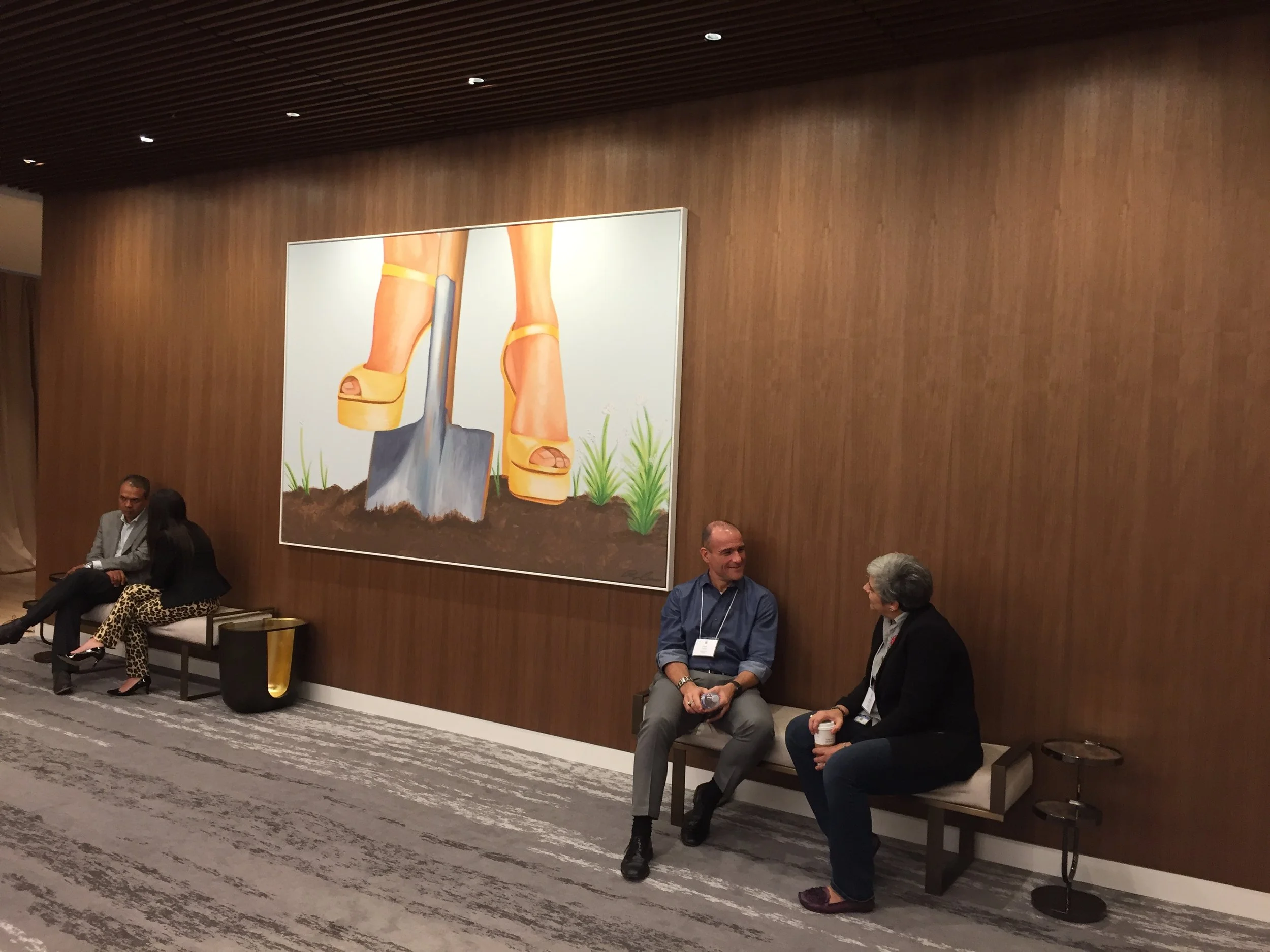 A group of people sitting and talking in a conference room with wood paneling on the walls and a large painting of high-heeled shoes and grass.
