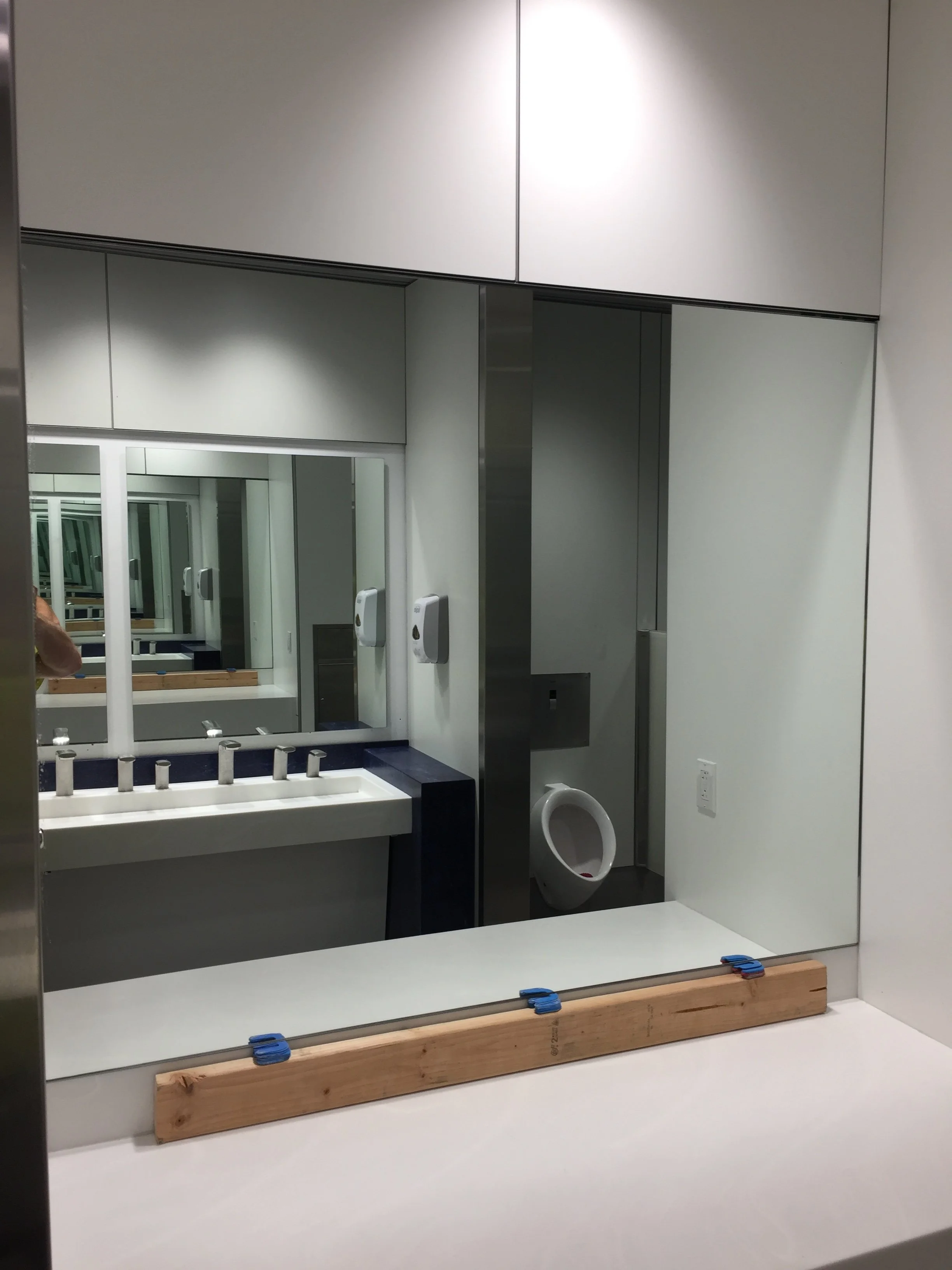 Reflection of a bathroom mirror showing a bathroom with a sink, a mirror, a toilet, white walls, and some wooden and blue clamps on the counter.