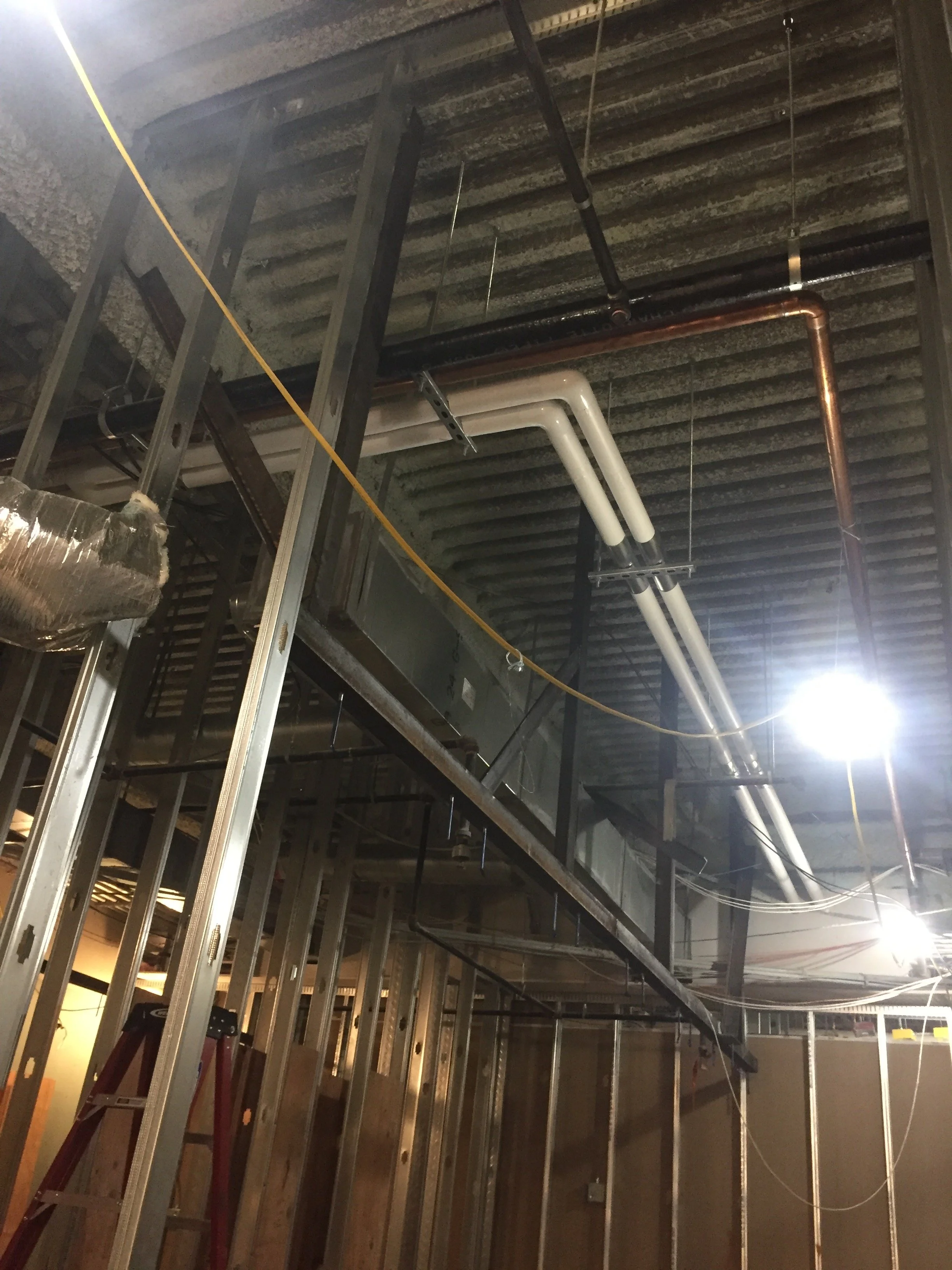 Construction site with exposed metal framing, pipes, and electrical wiring on unfinished ceiling.