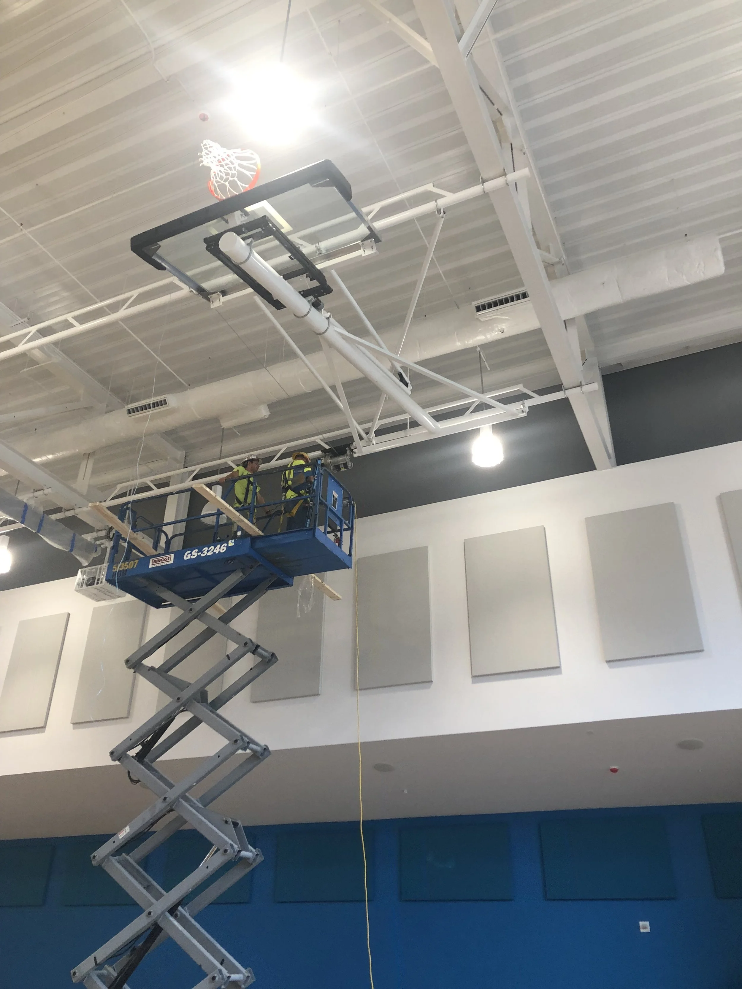 Workers on a blue scissor lift installing or maintaining ceiling fixtures in an interior space with a white ceiling and wall panels.