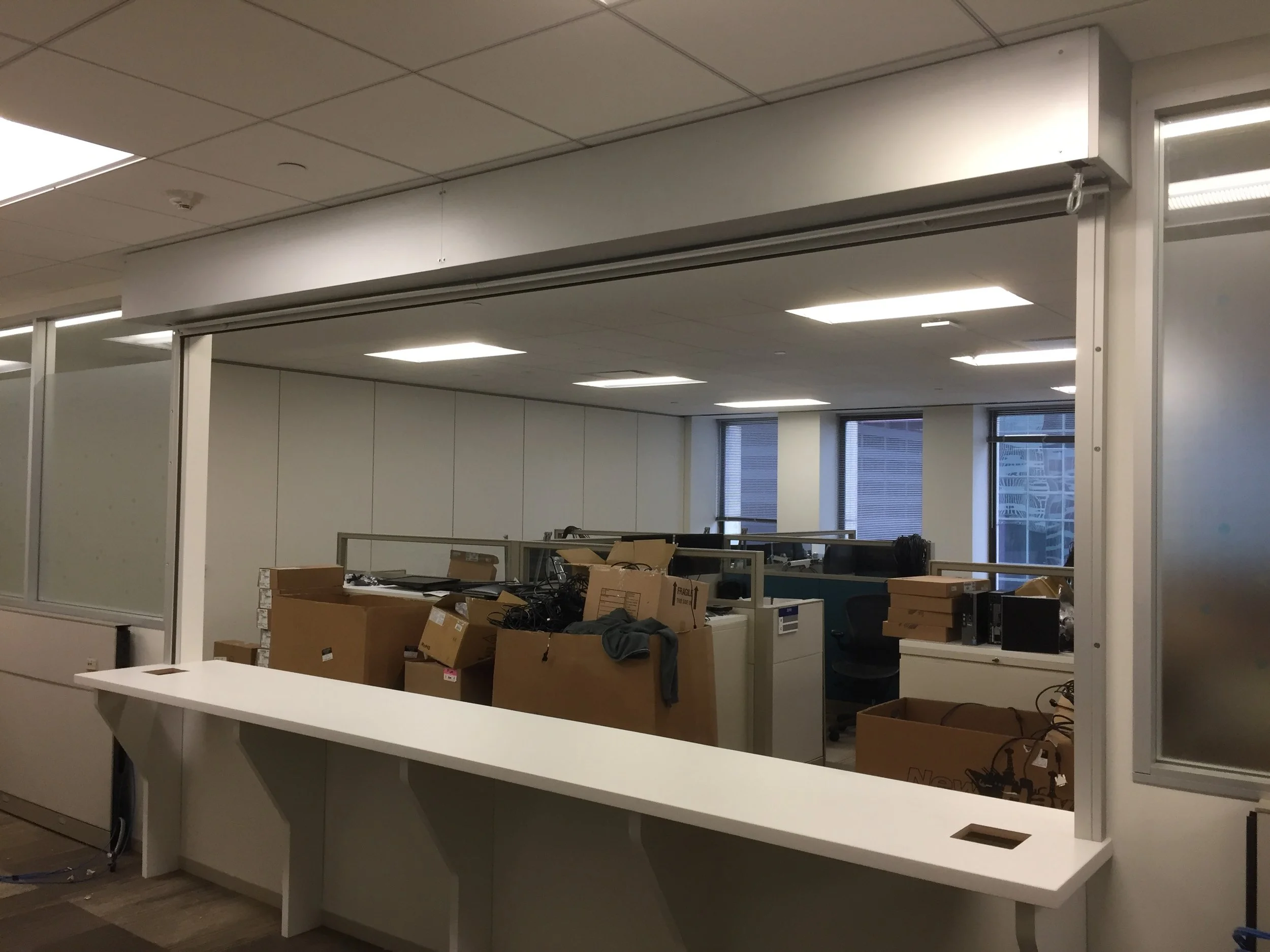 Empty office space with boxes and equipment on desks, white partition walls, large windows, and a white reception counter.