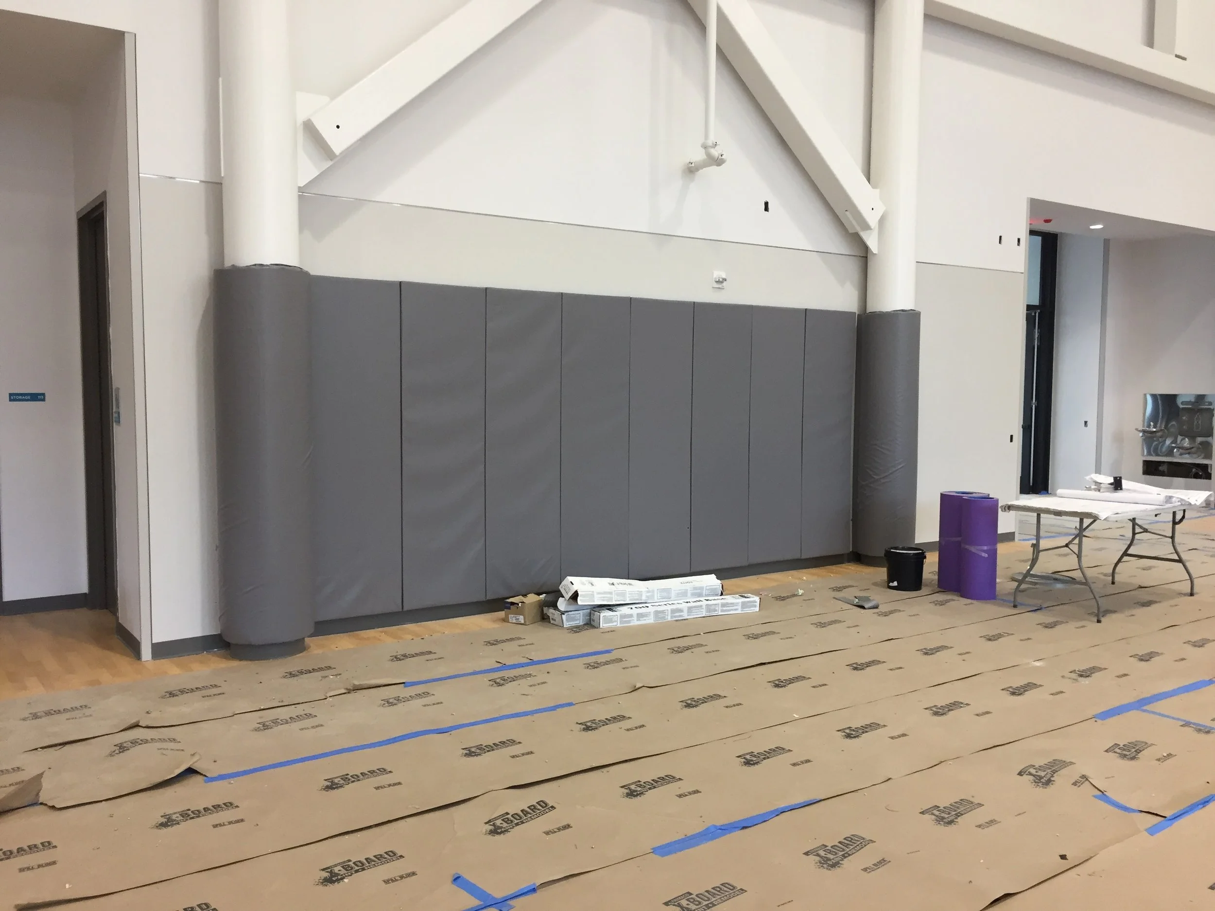 A construction or renovation site with a partially finished interior wall. The wall features vertical white beams and a section with gray padding or insulation. The floor is covered temporarily with printed cardboard and blue tape, and there are cons