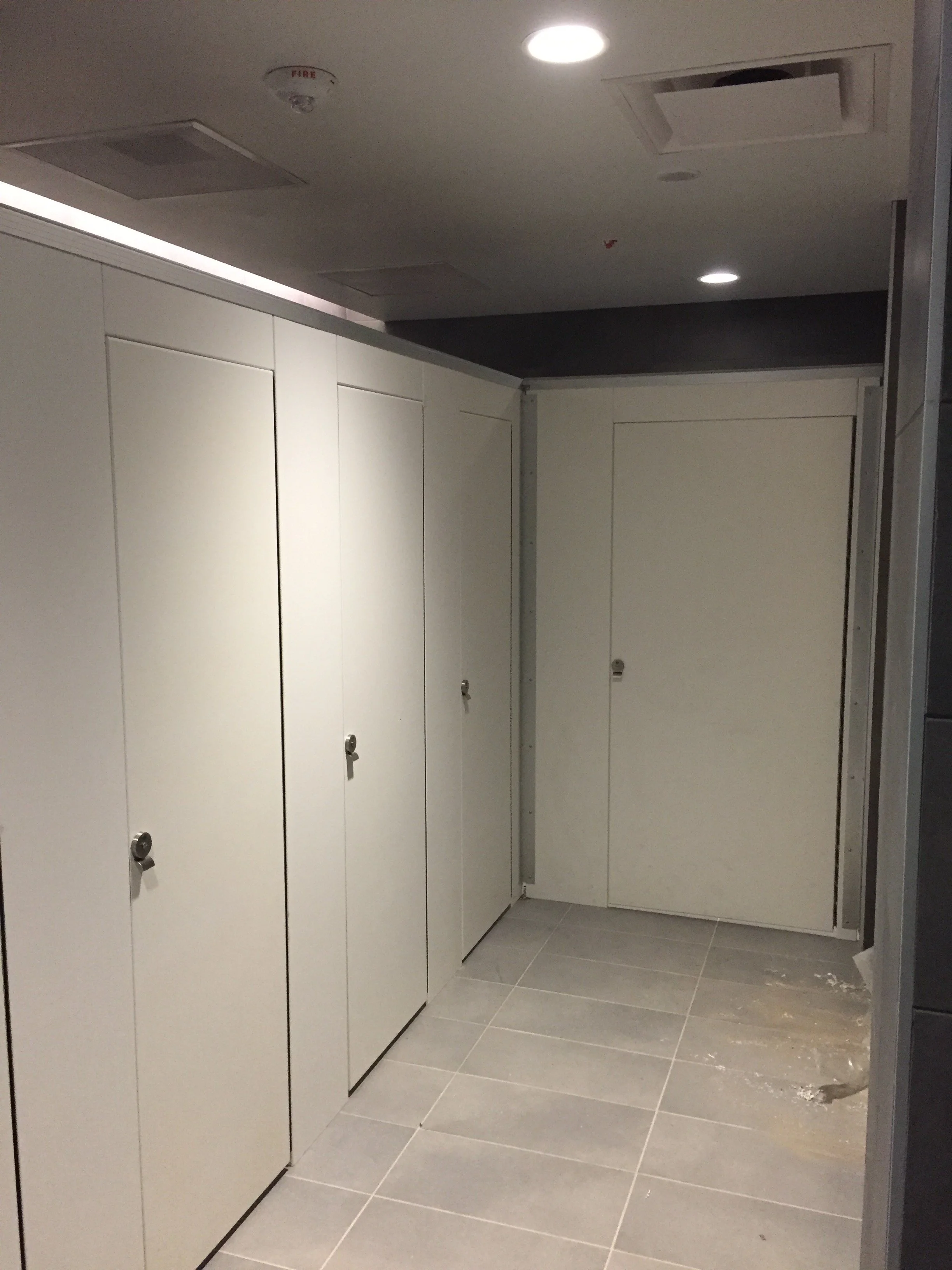 Public restroom with white stalls, closed doors, silver locks, beige tiled floor, ceiling lights, and a smoke detector.