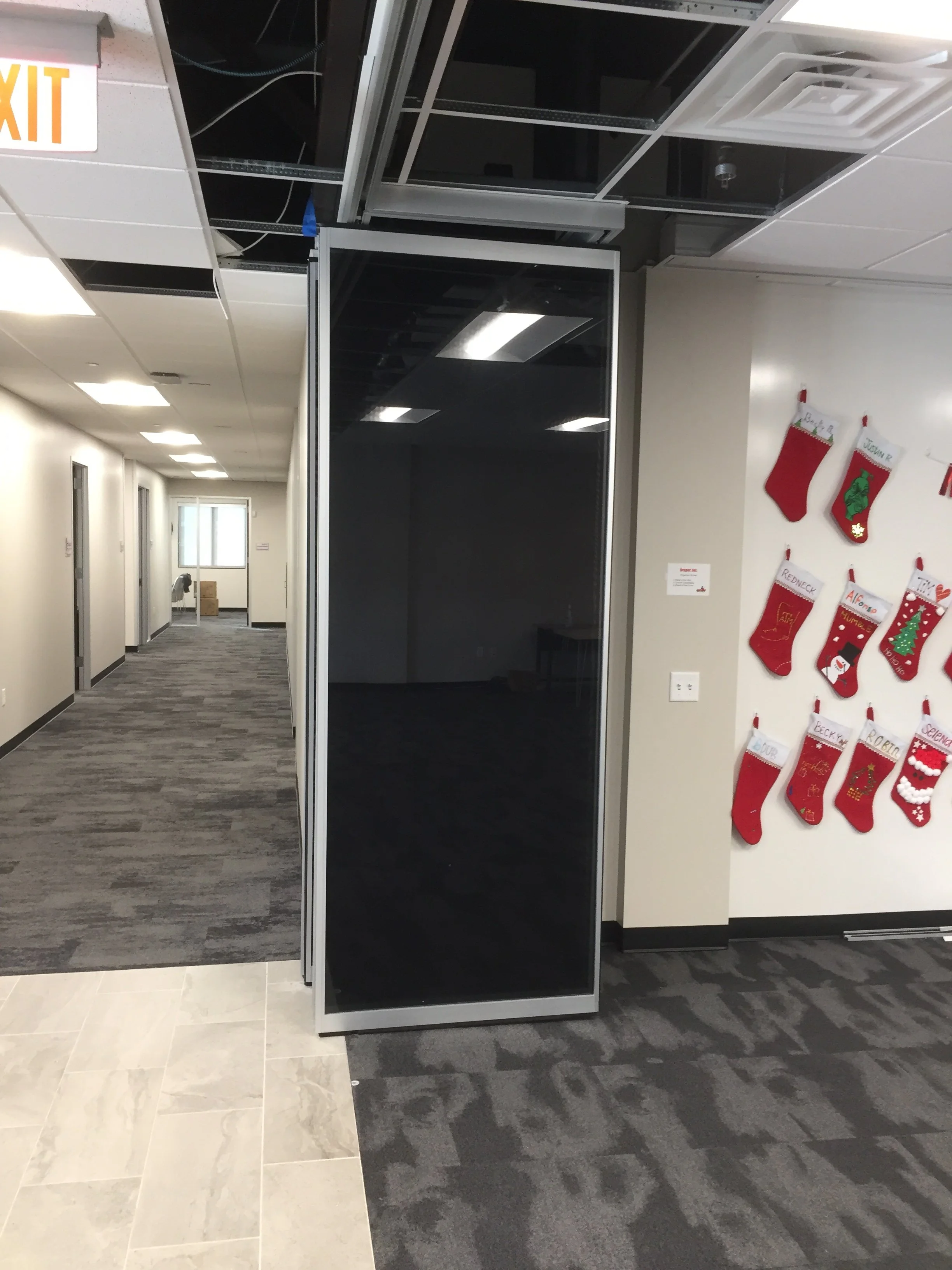 Empty hallway with a suspended ceiling, gray and beige tile flooring, and several doors on the left. On the right, there is a Christmas stocking display on the wall with red stockings decorated with holiday motifs and names. A glass and metal display