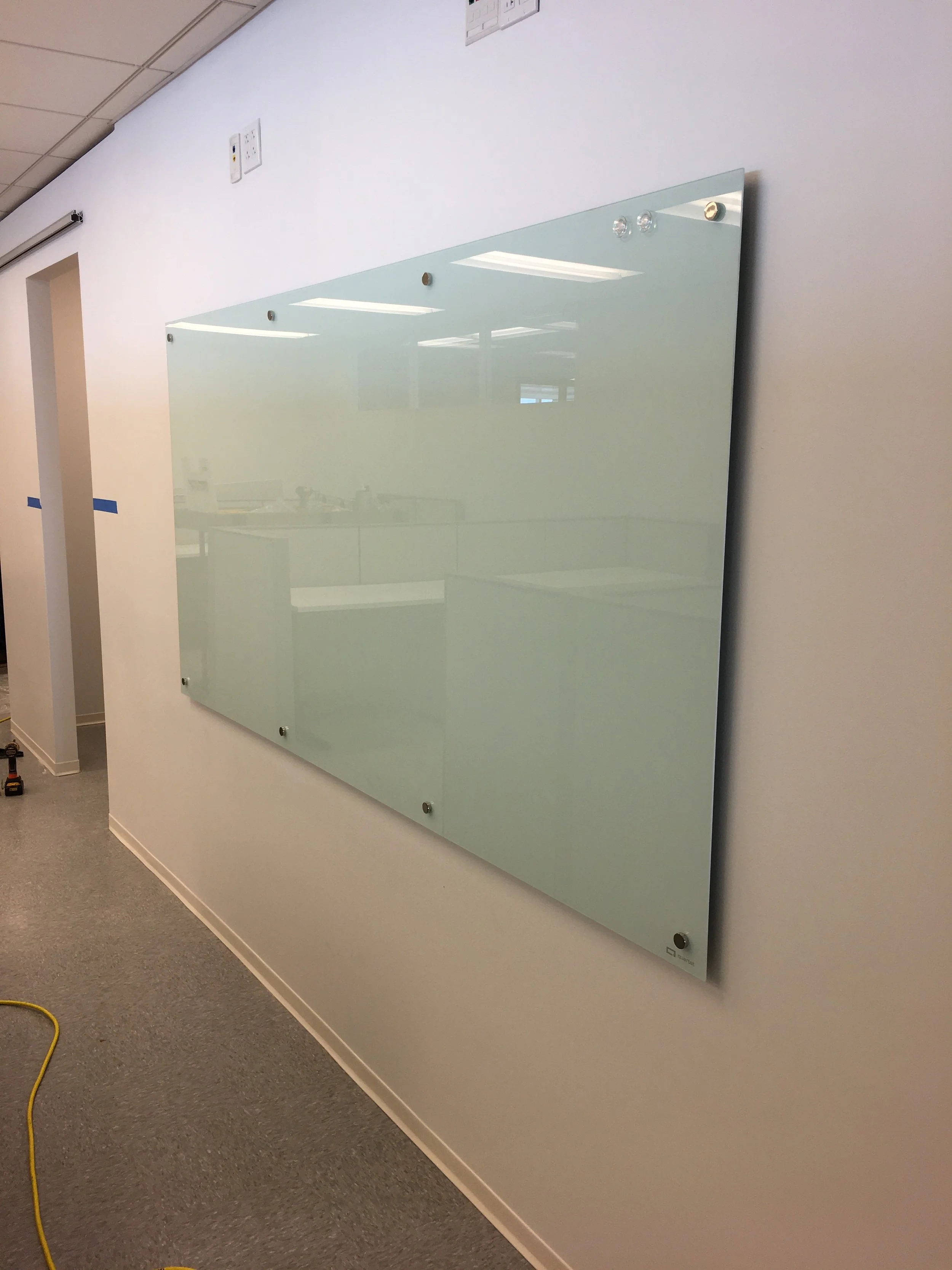 A frosted glass whiteboard mounted on a white wall in an office with electrical outlets and ceiling lights reflected in the glass.