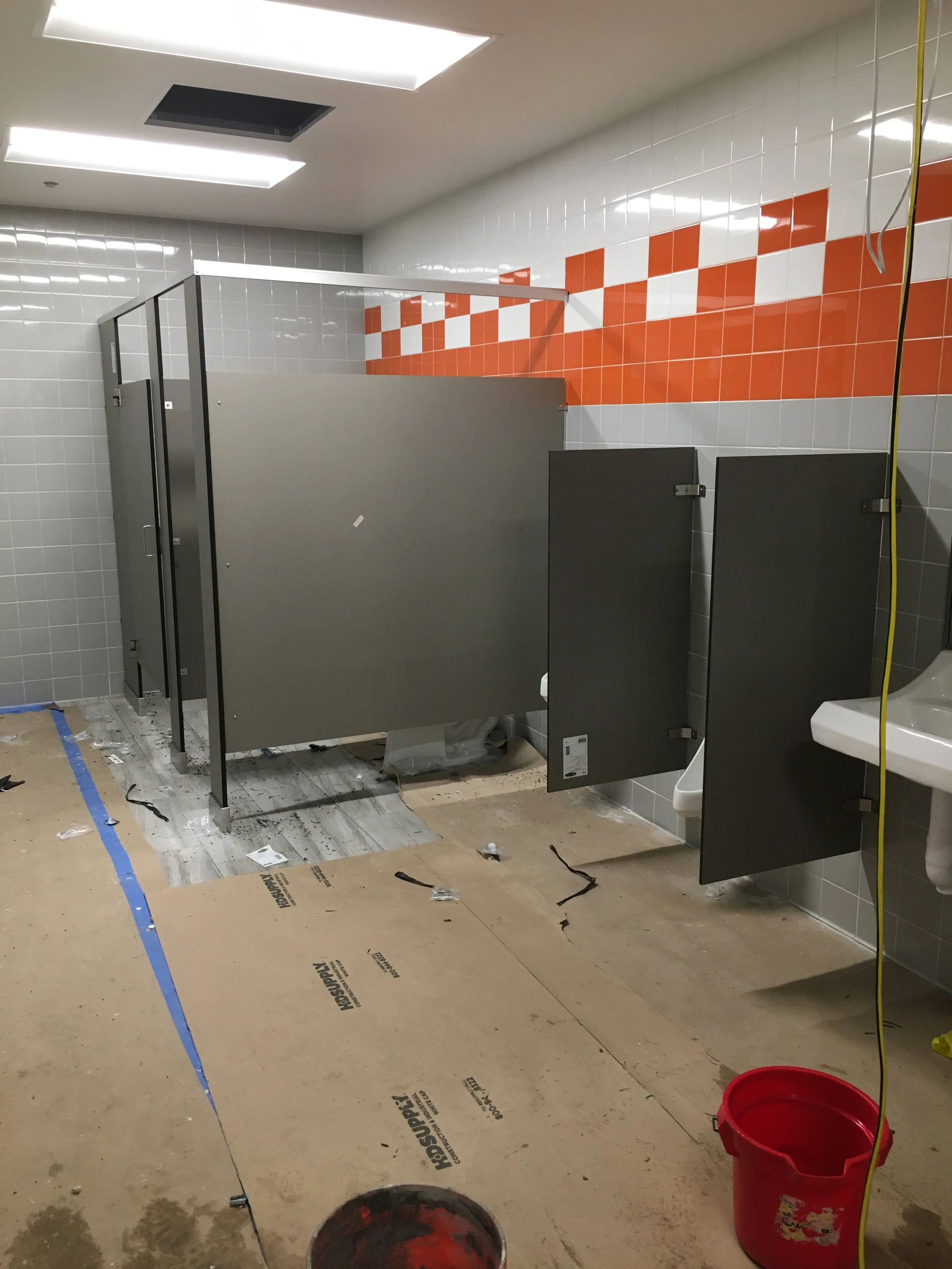 Bathroom under construction with partially installed stalls, tiled walls, sawdust on the floor, a red bucket, and construction tools.