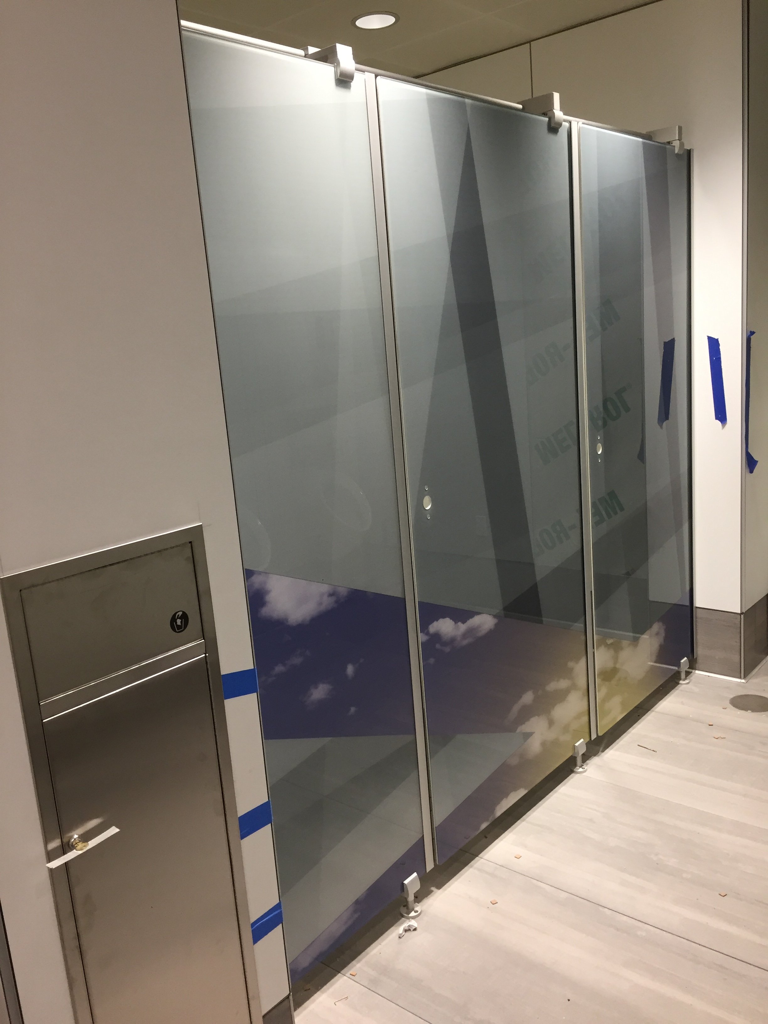 Public restroom with multiple toilet stalls, one of which has a door decorated with a sky and clouds image. The stalls are metallic with locks, and the floor is light-colored wood or laminate. There is a small trash bin near the stalls.