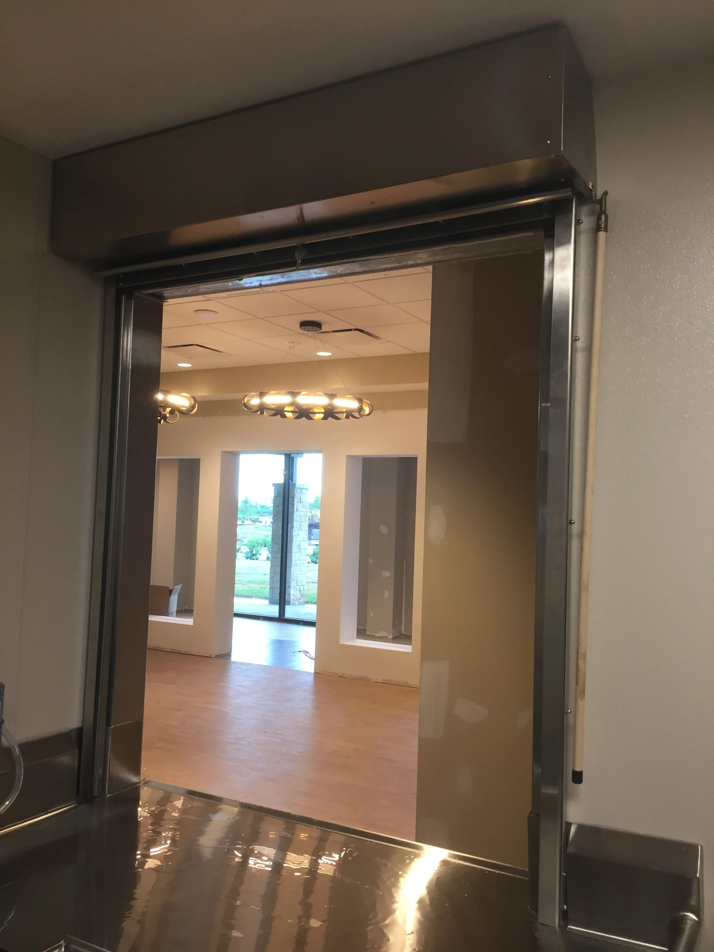 An elevator door opening into a room with wooden flooring, beige walls, and modern ceiling lights. Large windows reveal a view outside.