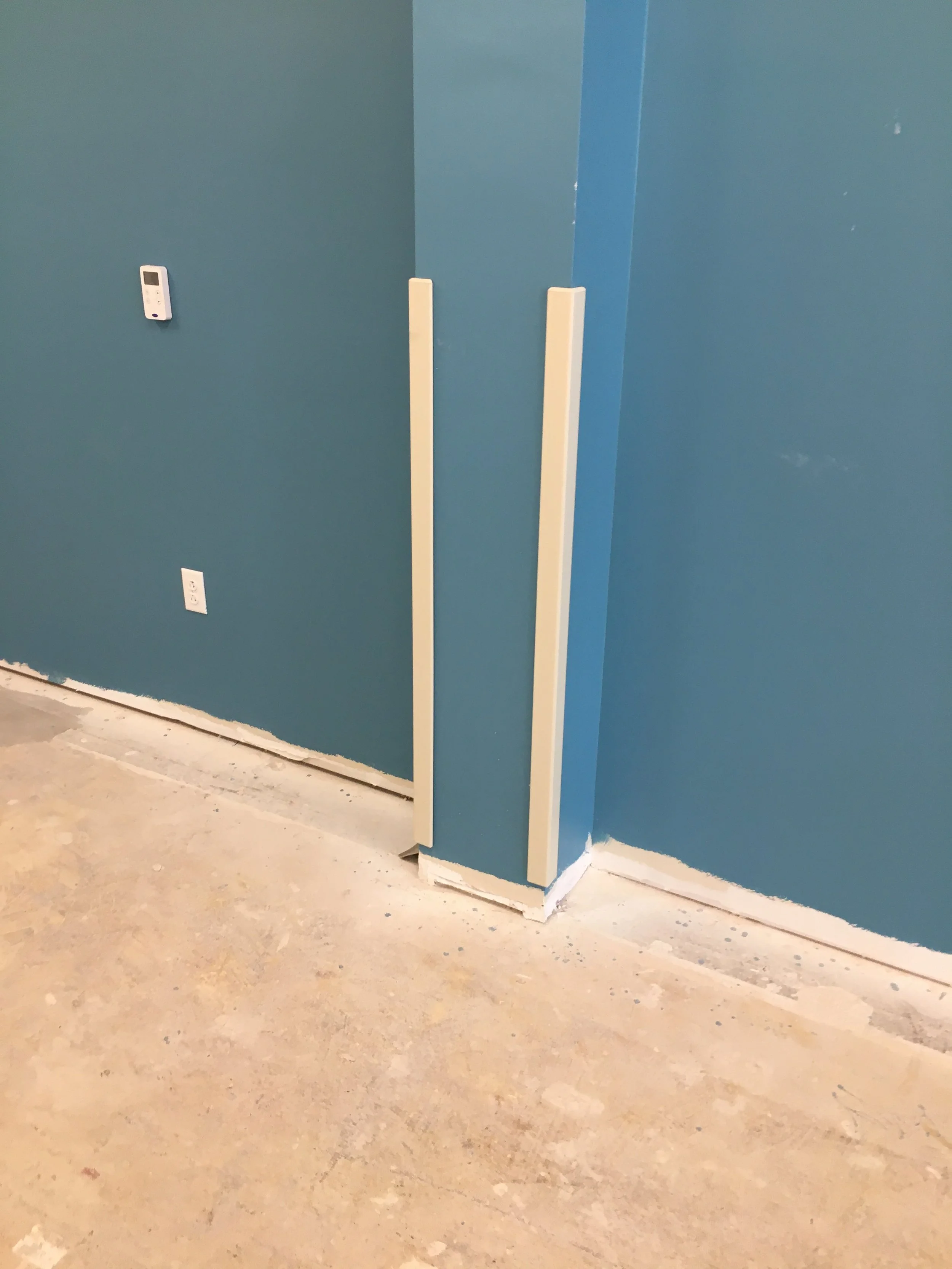 Interior wall with blue paint, partially painted, with baseboards and electrical outlets visible, and an unfinished floor.