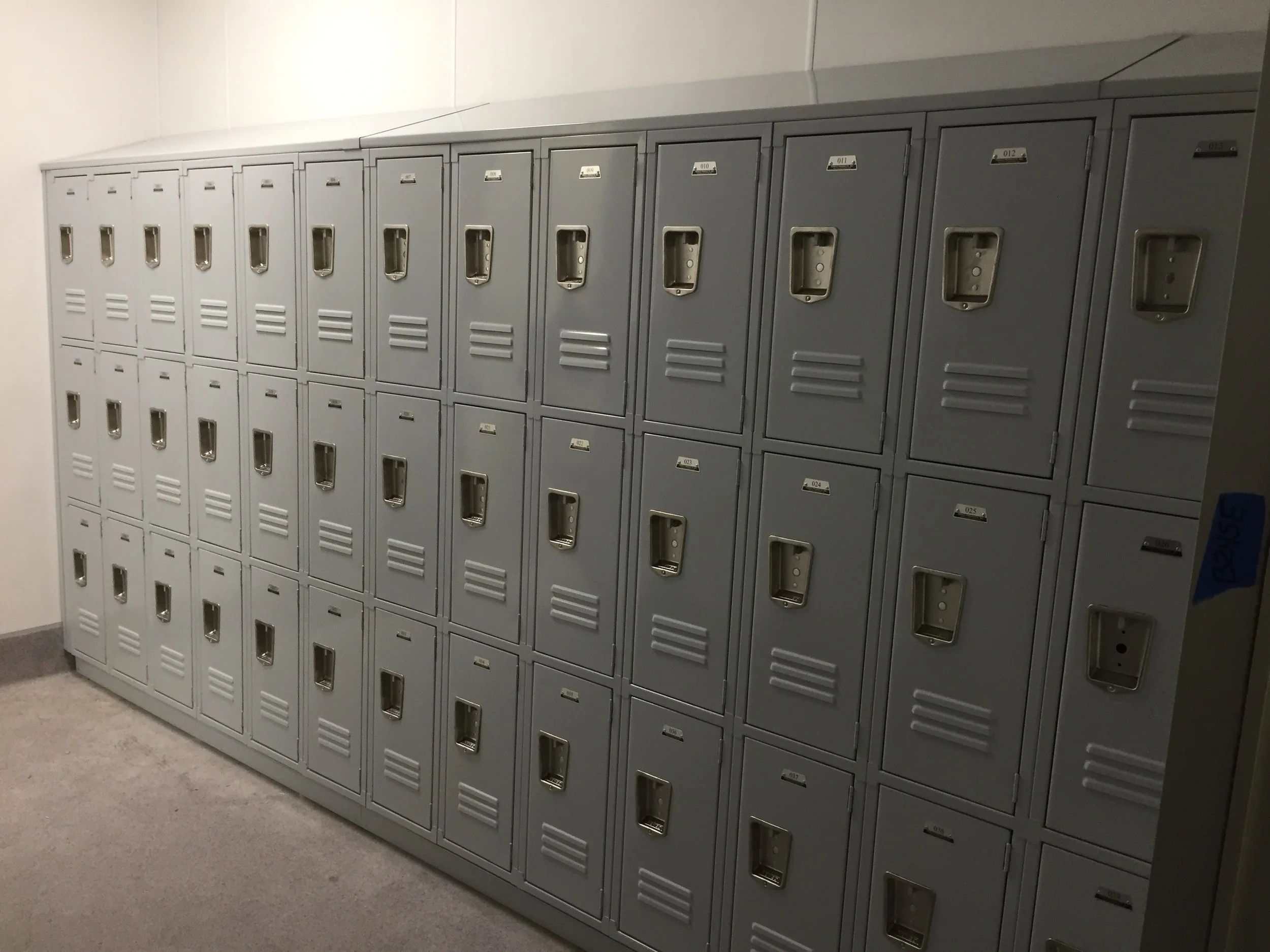 Row of grey metal lockers with vented doors, numbered labels, and open slots in a school or gym locker room, with a grey floor and plain wall.