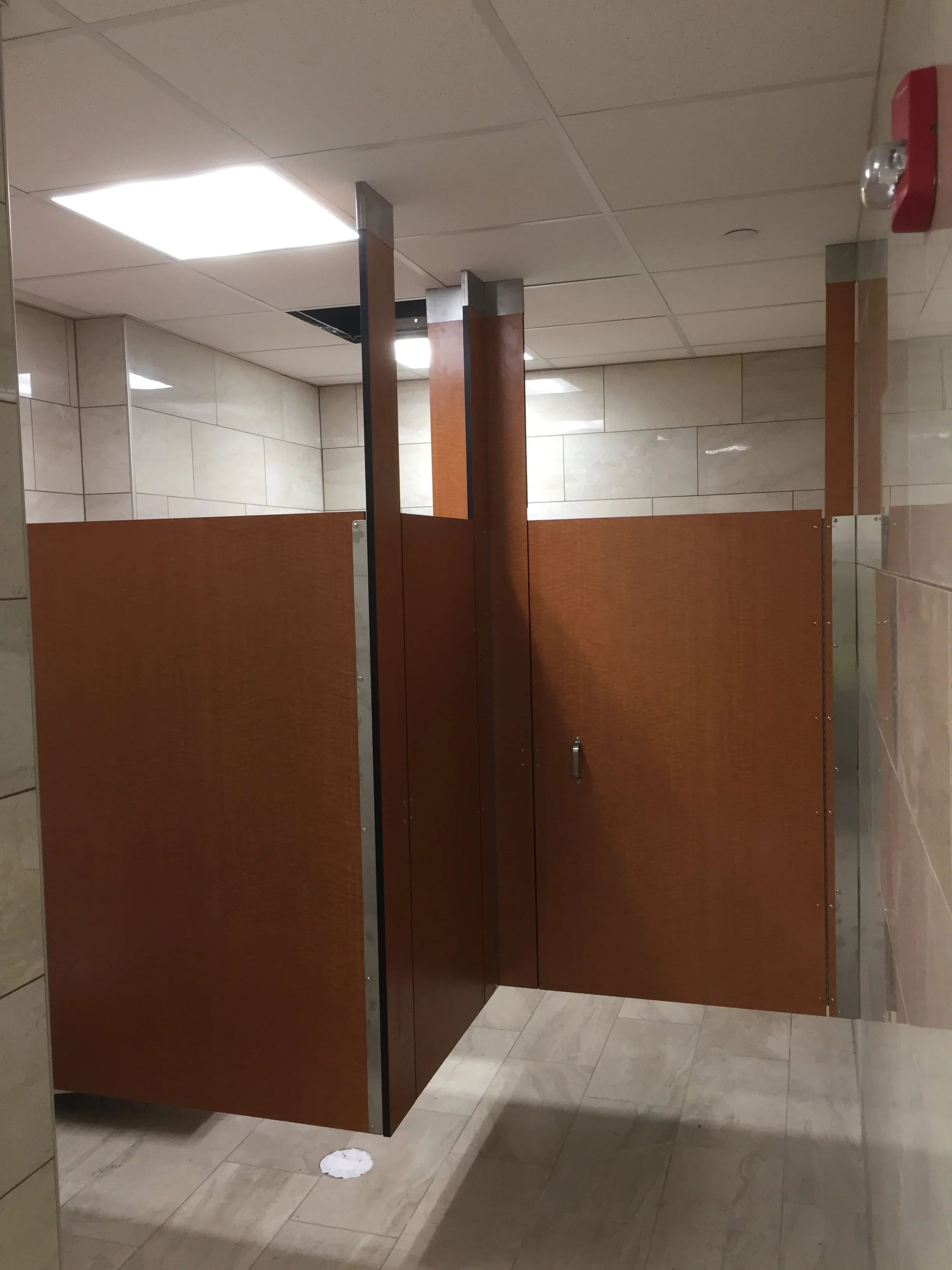 A public restroom stall with a wooden door and walls, beige tiled floor, and white tiled walls. A small white paper towel or tissue is seen on the floor near the stall.