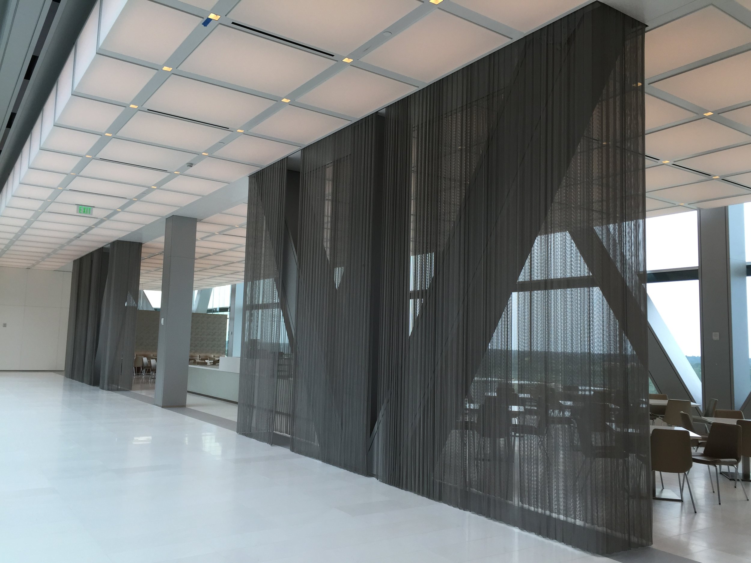 Modern interior with a high ceiling, geometric design, large glass windows, metal mesh partitions, seating area, and an exit sign.