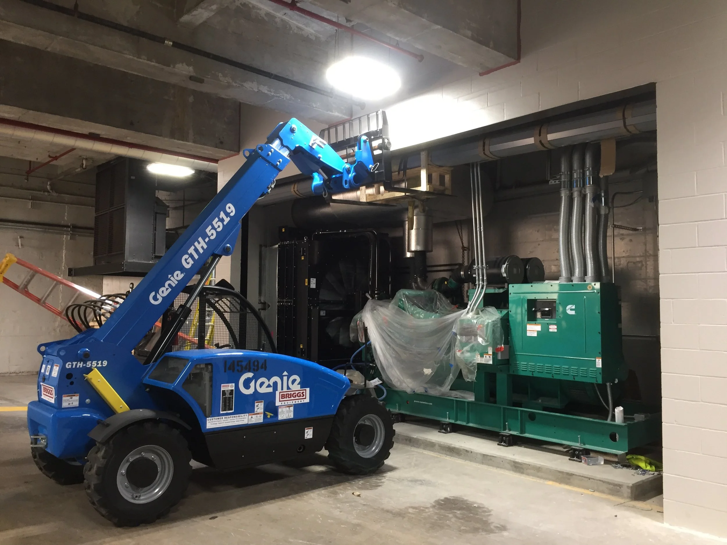 Blue Genie GTH-5519 compact utility loader lifting and transporting a green equipment cart in an underground parking garage with exposed ceiling pipes and concrete walls.