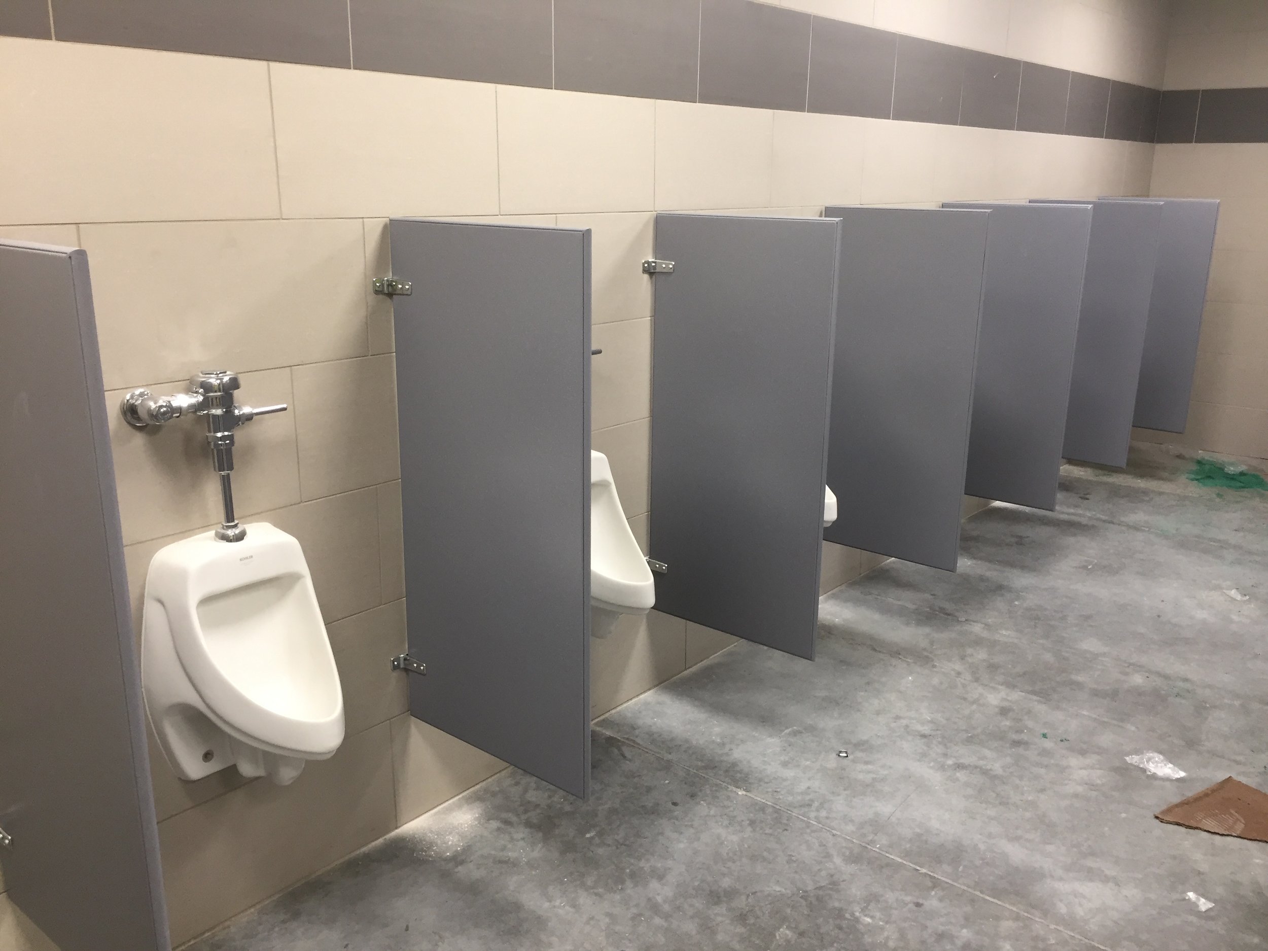 A public restroom with three urinals separated by gray partitions, beige tiled walls, and a concrete floor with some trash and a mat.