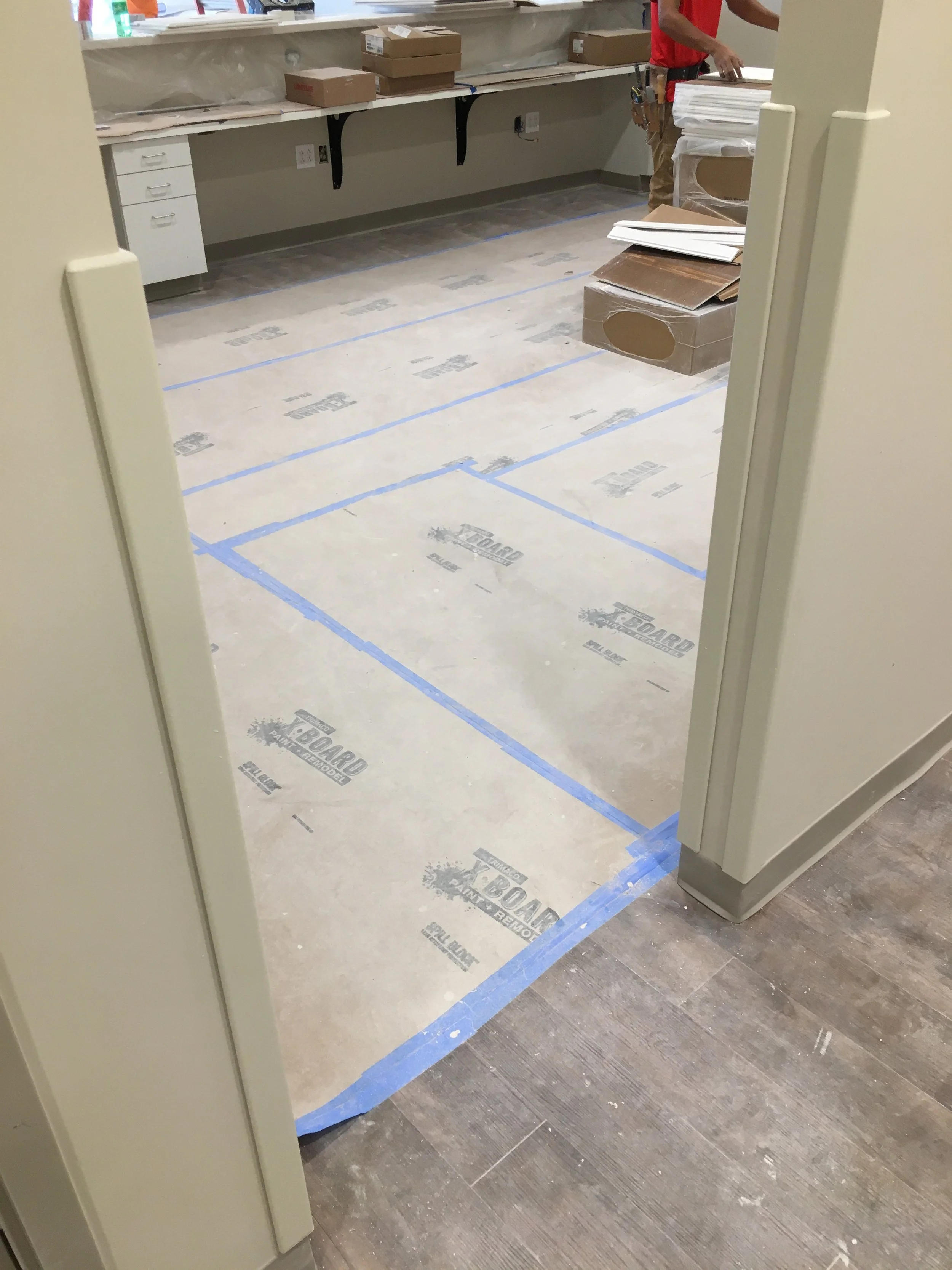 Construction or renovation site with floor covered in protective paper and blue tape, partially visible worker in red shirt and tool belt, boxes and materials on counter and floor.