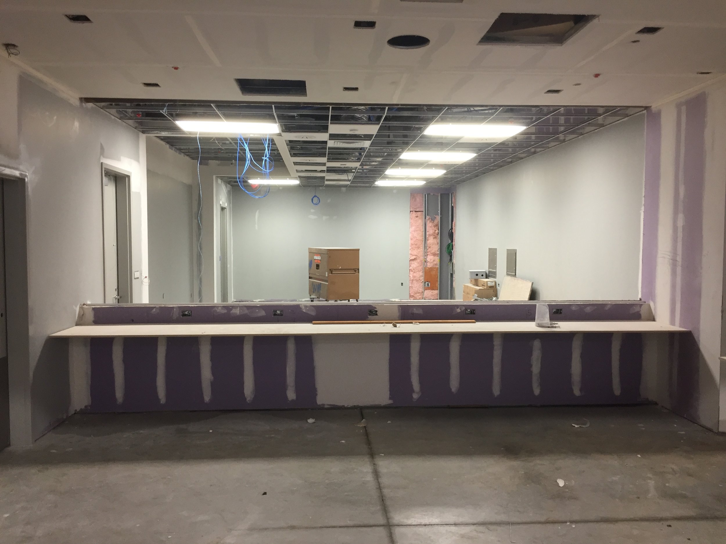 Construction area with unfinished ceiling panels, exposed wires, and construction materials, viewed from a lower angle in an interior space.