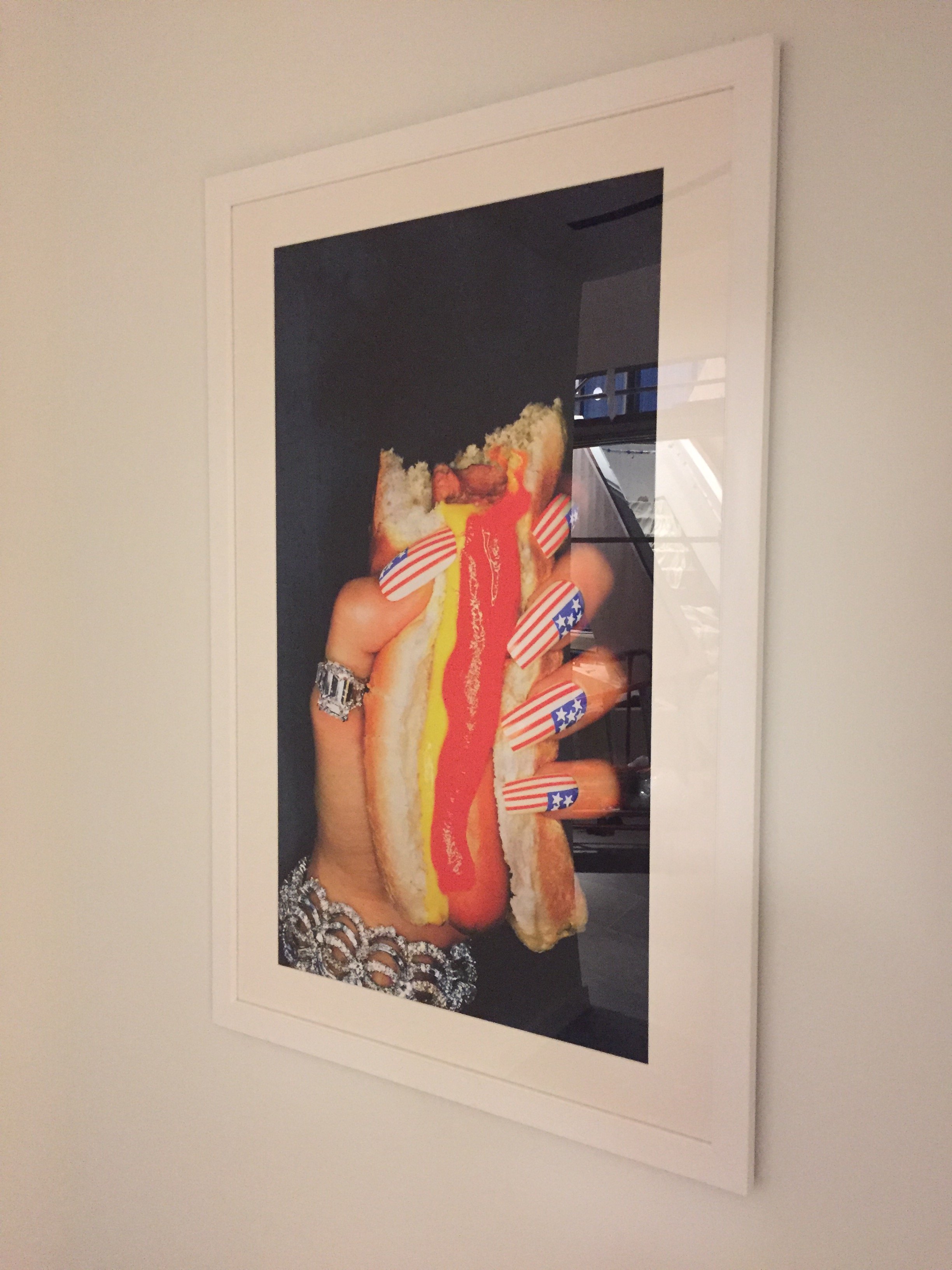 Photograph of a hand holding a hot dog with mustard, ketchup, and American flag-themed nails. The photo is framed and displayed on a wall.