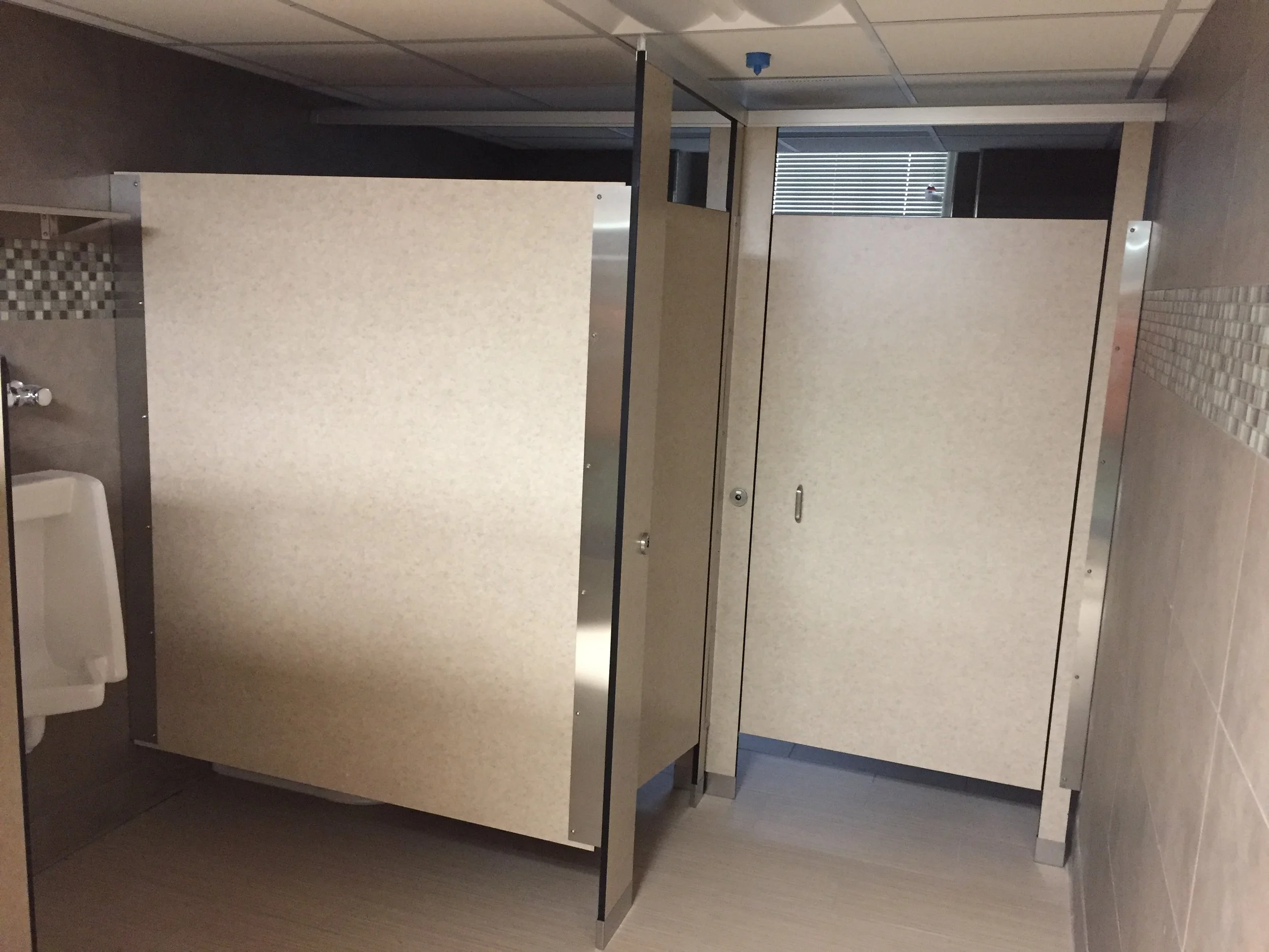 Public restroom stalls with beige doors, partially open stall door with a paper towel dispenser visible, tile flooring, and window blinds in the background.